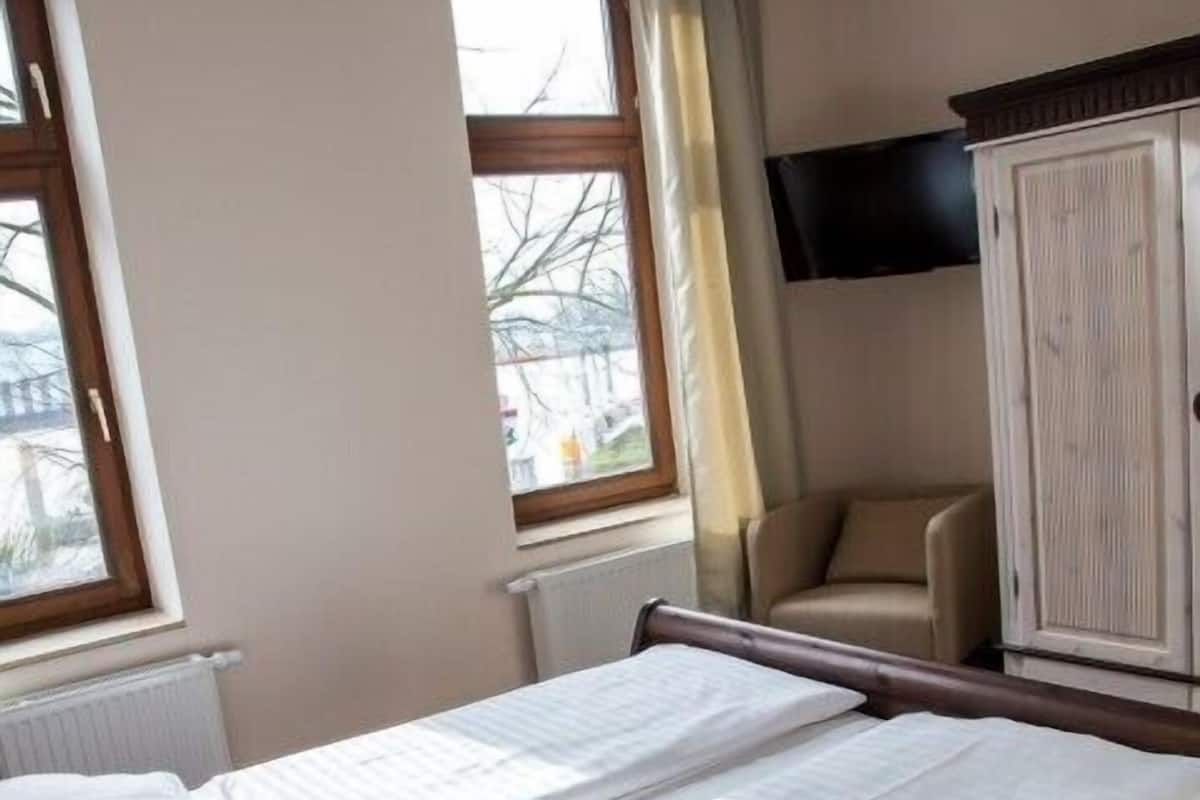 A room with two single beds, a wooden wardrobe, a television mounted on the wall, and a window with a view of the outside.