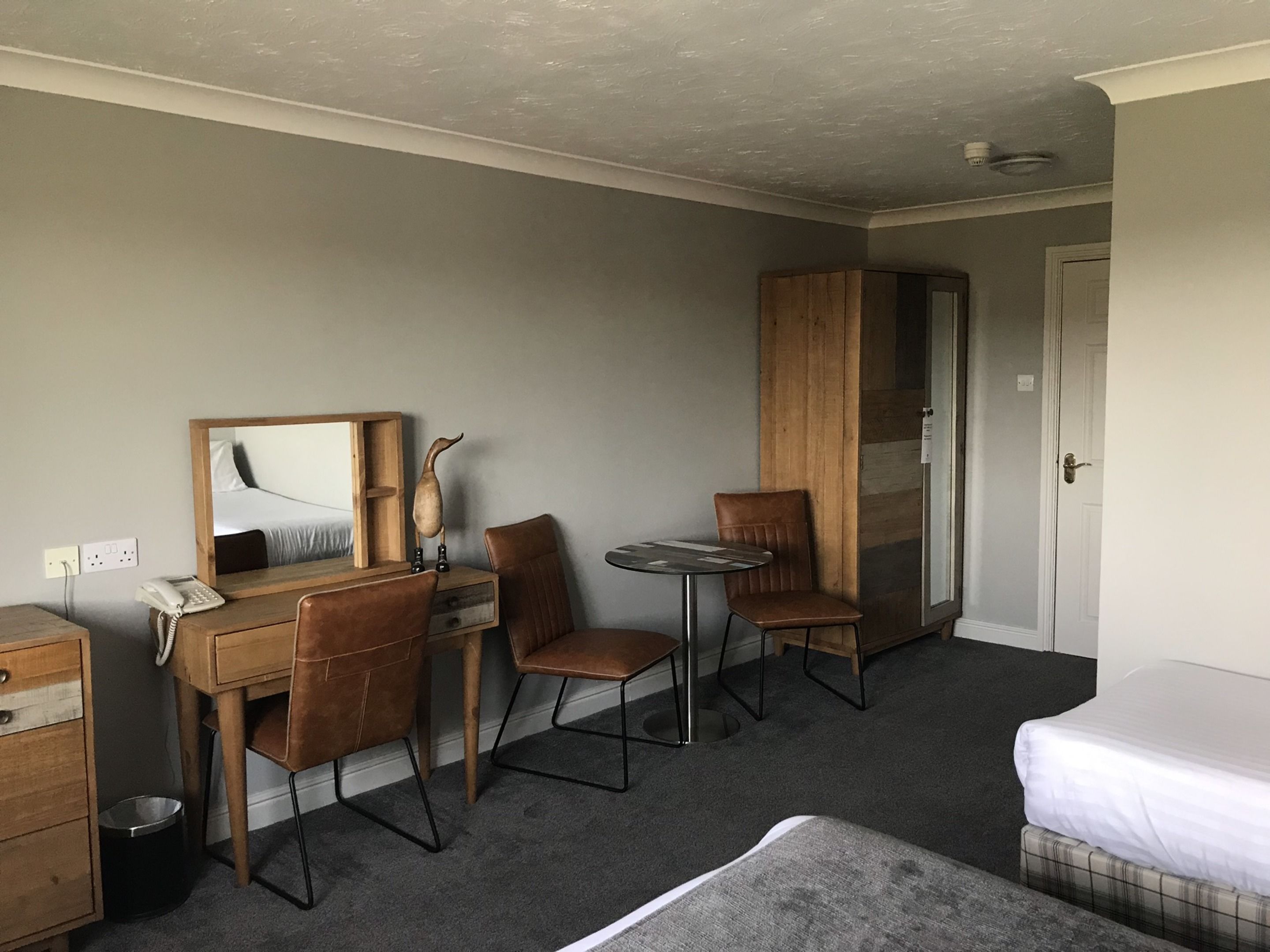 Habitación de hotel con una cama, un escritorio con silla, una mesita con otra silla, un armario y un espejo en la pared.
