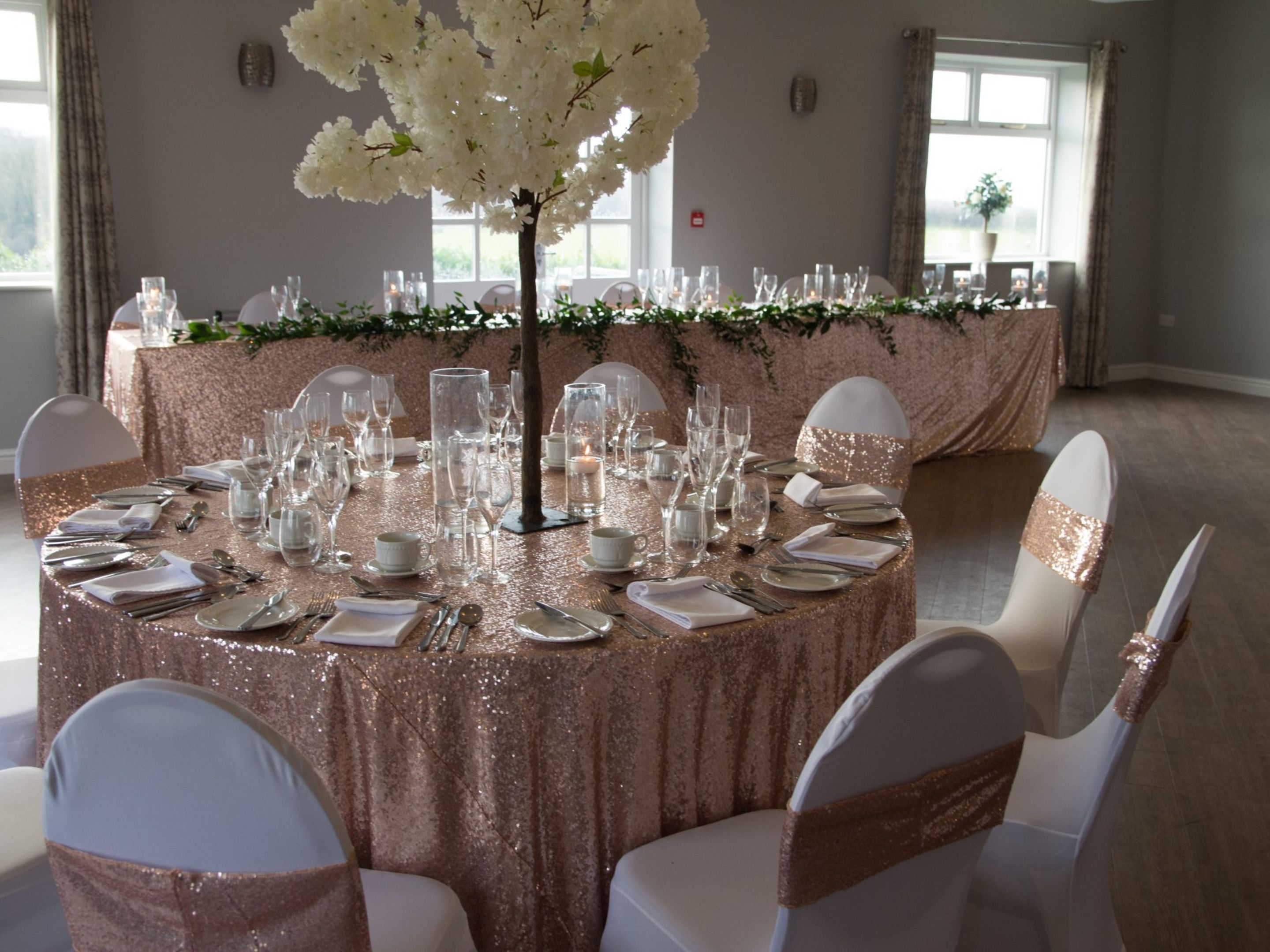 Une table ronde recouverte d’une nappe scintillante, entourée de chaises blanches ornées de rubans rose doré, préparée pour un événement formel.
