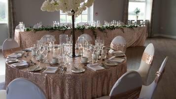 Una mesa redonda con mantel brillante, rodeada de sillas blancas con cintas doradas, lista para un evento formal.