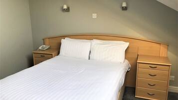 A neatly made bed with white linens in a room with a wooden headboard. There are two bedside tables, each with a lamp and a phone. The room has a simple, clean design with light-colored walls and a carpeted floor.