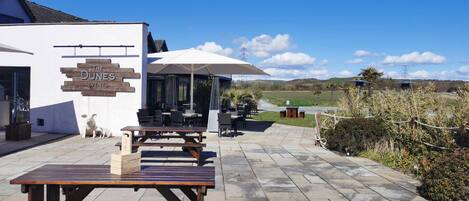 An outdoor seating area with tables and chairs, a white building with 'The Dunes Events' sign, and a view of the countryside.