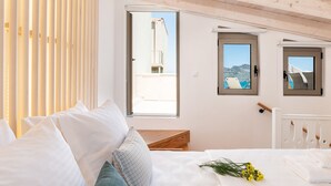 A bedroom with a bed, white pillows, a wooden bench, and a window with a view of a building and mountains. - Sea Front Villas (Chania)