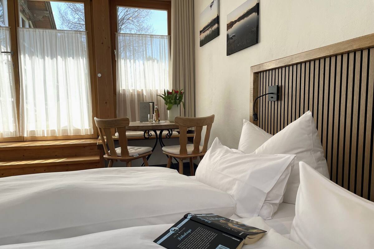 A hotel room with a bed, a table with chairs, a window with curtains, and a wall-mounted lamp.