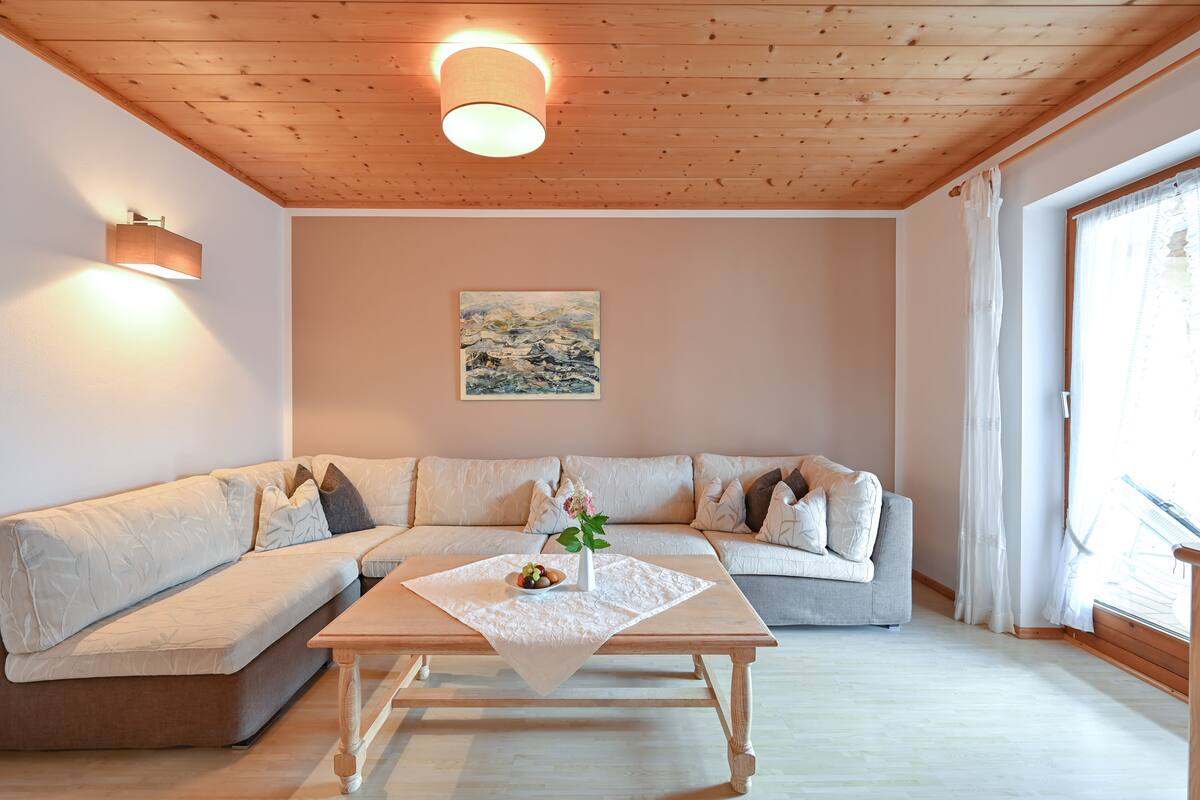 A modern living room with a wooden ceiling, a beige sofa, a wooden coffee table with a vase and fruit bowl, and a painting on the wall.