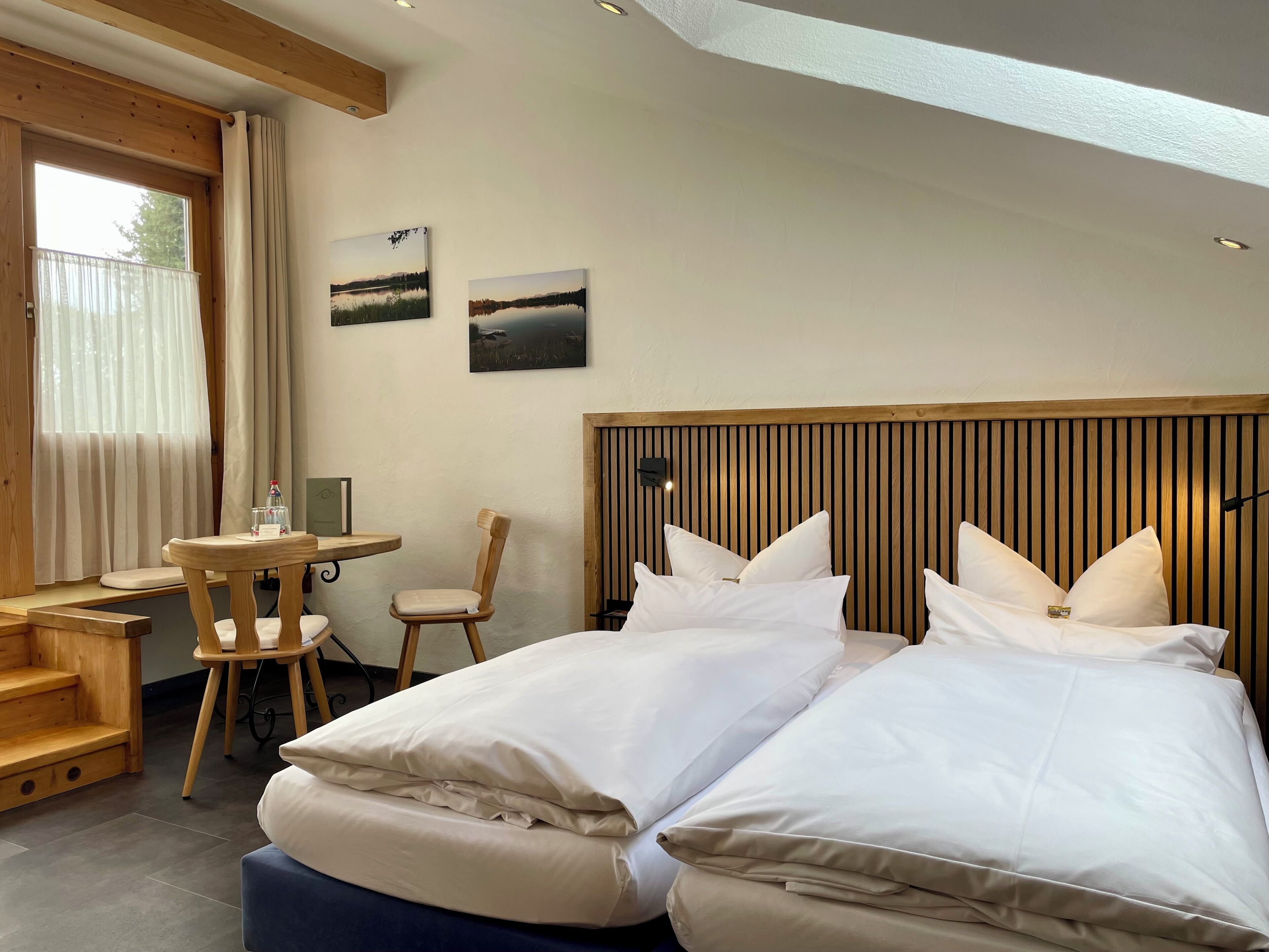 A hotel room with a wooden headboard, two beds with white bedding, a small wooden table with a chair, and two framed pictures on the wall.