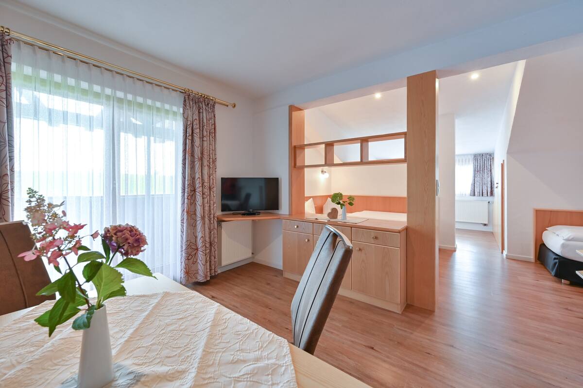 A modern hotel room with a wooden floor, a dining area with a table and chairs, a TV, a small kitchen area, and a separate bedroom.