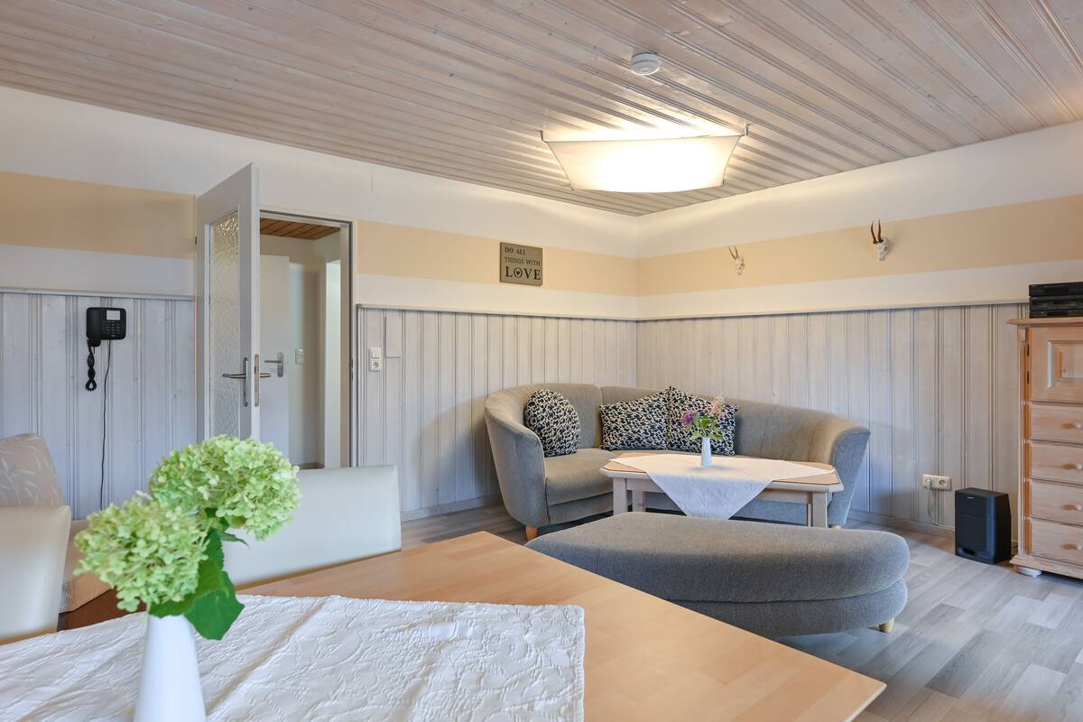 A cozy room with a wooden ceiling, a grey sofa, a small table, and a white vase with flowers.