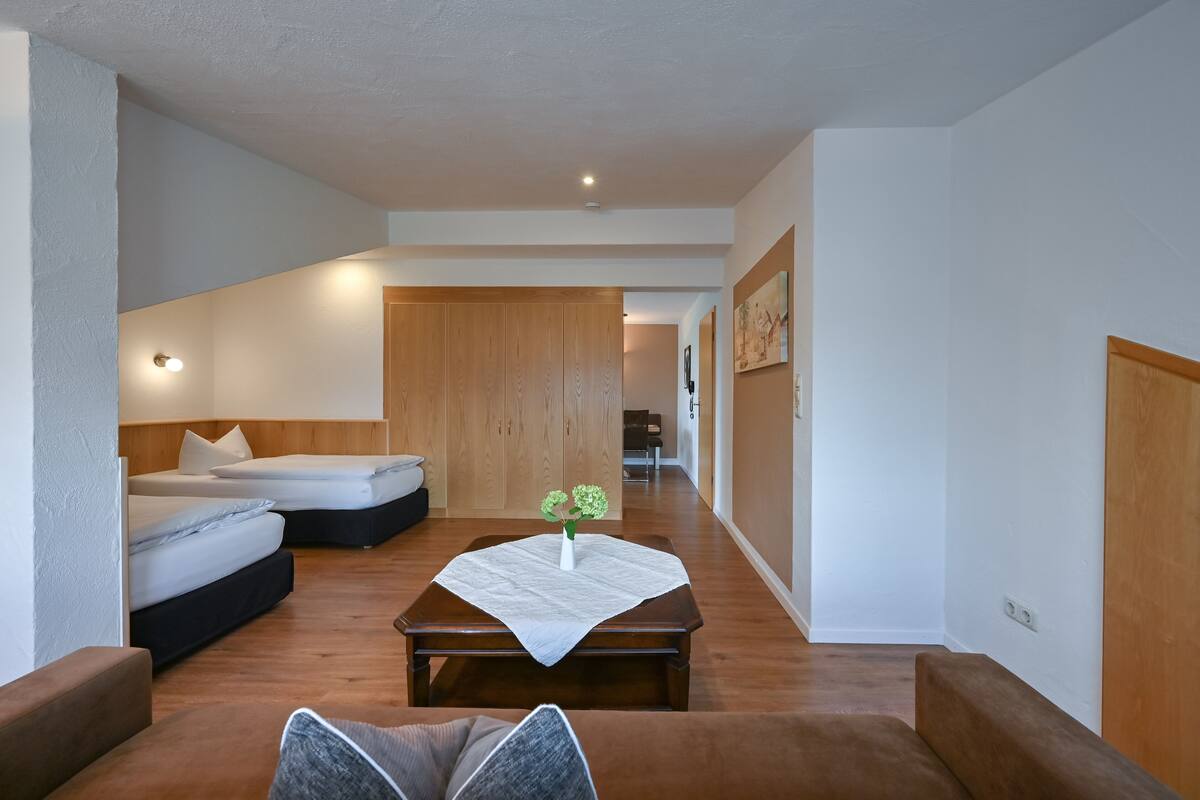 A modern hotel room with a wooden floor, two beds, a small table with a vase, and a hallway leading to another room.