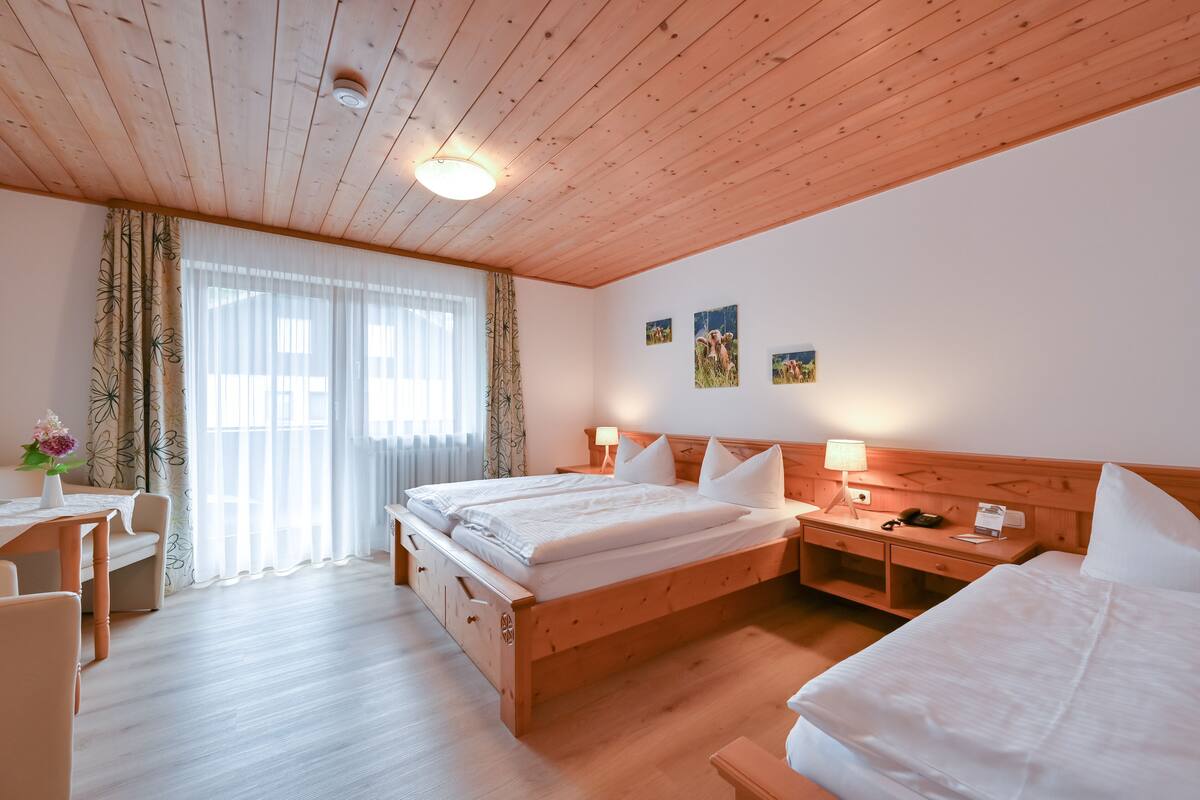 A hotel room with a wooden ceiling, a large bed with white linens, a desk with a lamp, and a window with sheer curtains.