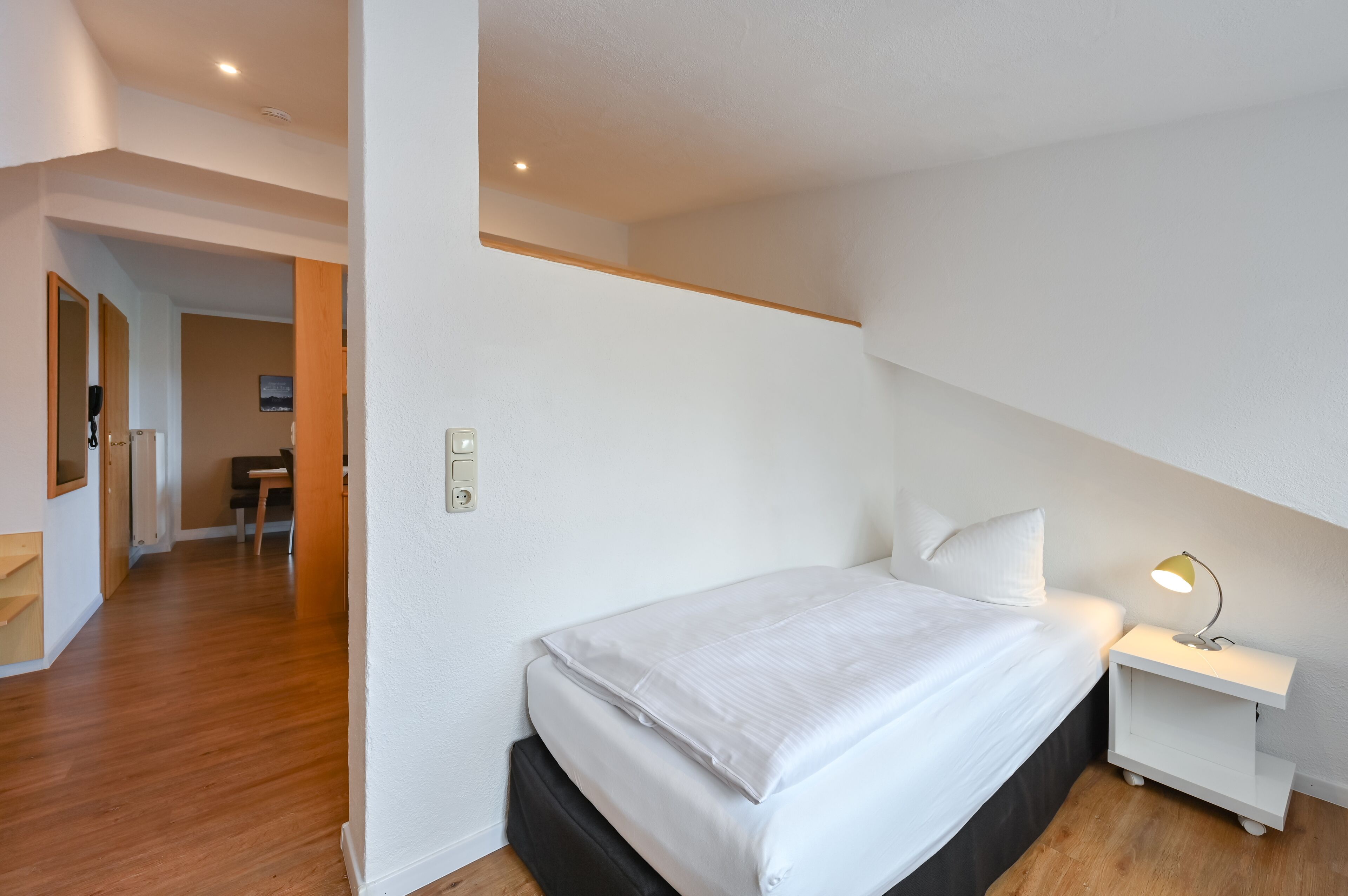 A bedroom with a bed, bedside table, and a lamp. There is a door leading to another room.