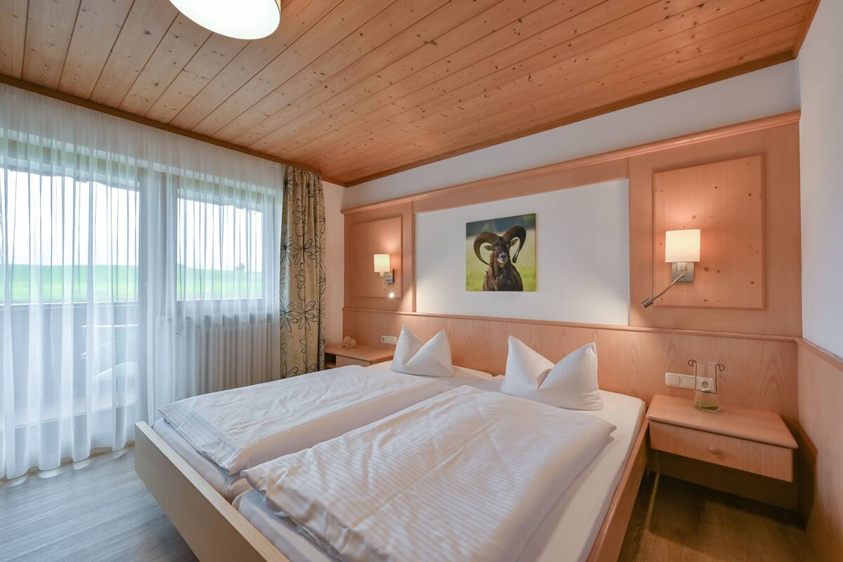 A double bed with white linens, a wooden headboard, bedside lamps, and a painting of a ram.