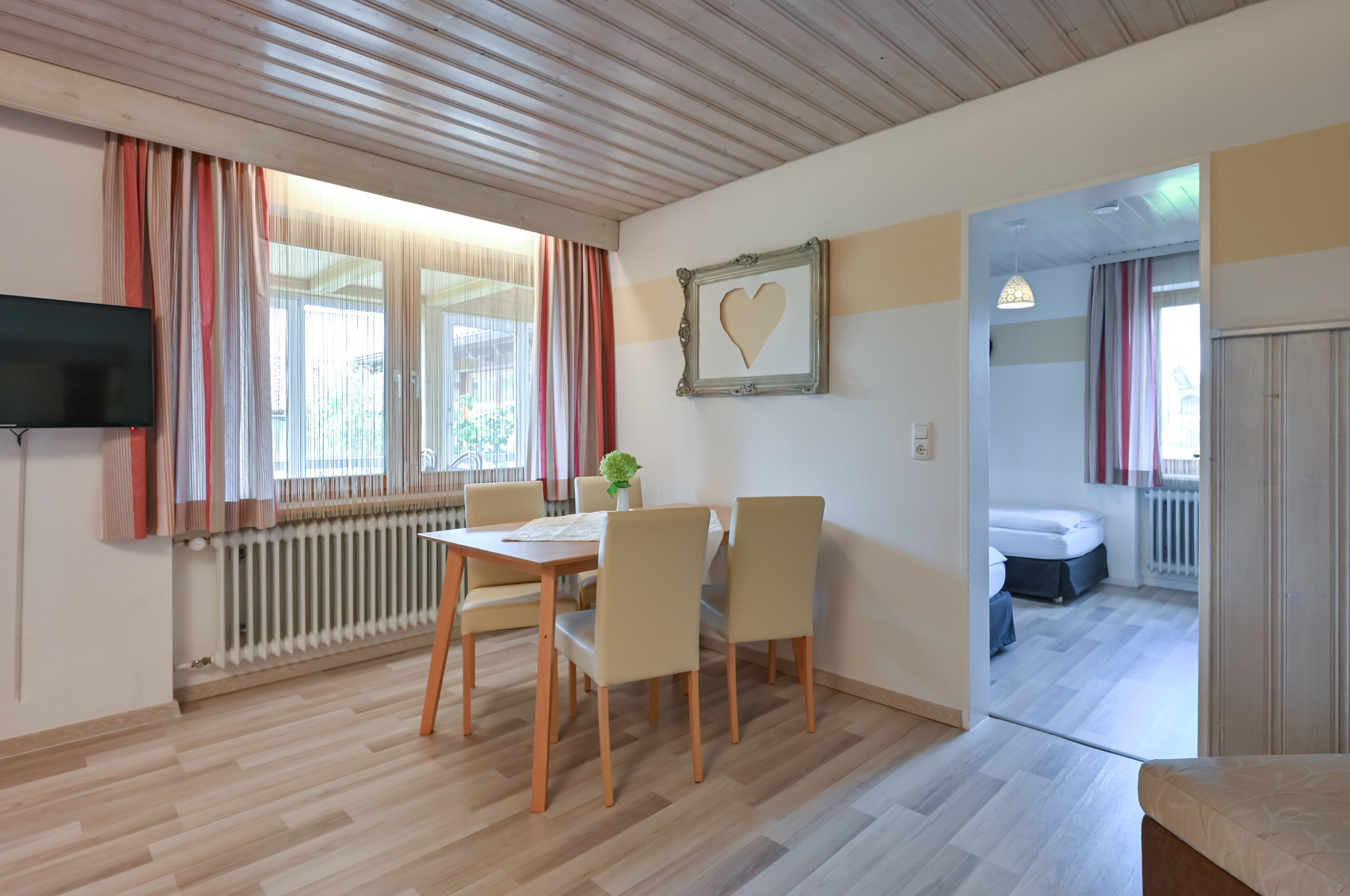Huoneessa on ruokapöytä, tuoleja, televisio, patteri ja sydämenmuotoinen valokuvakehys seinällä.