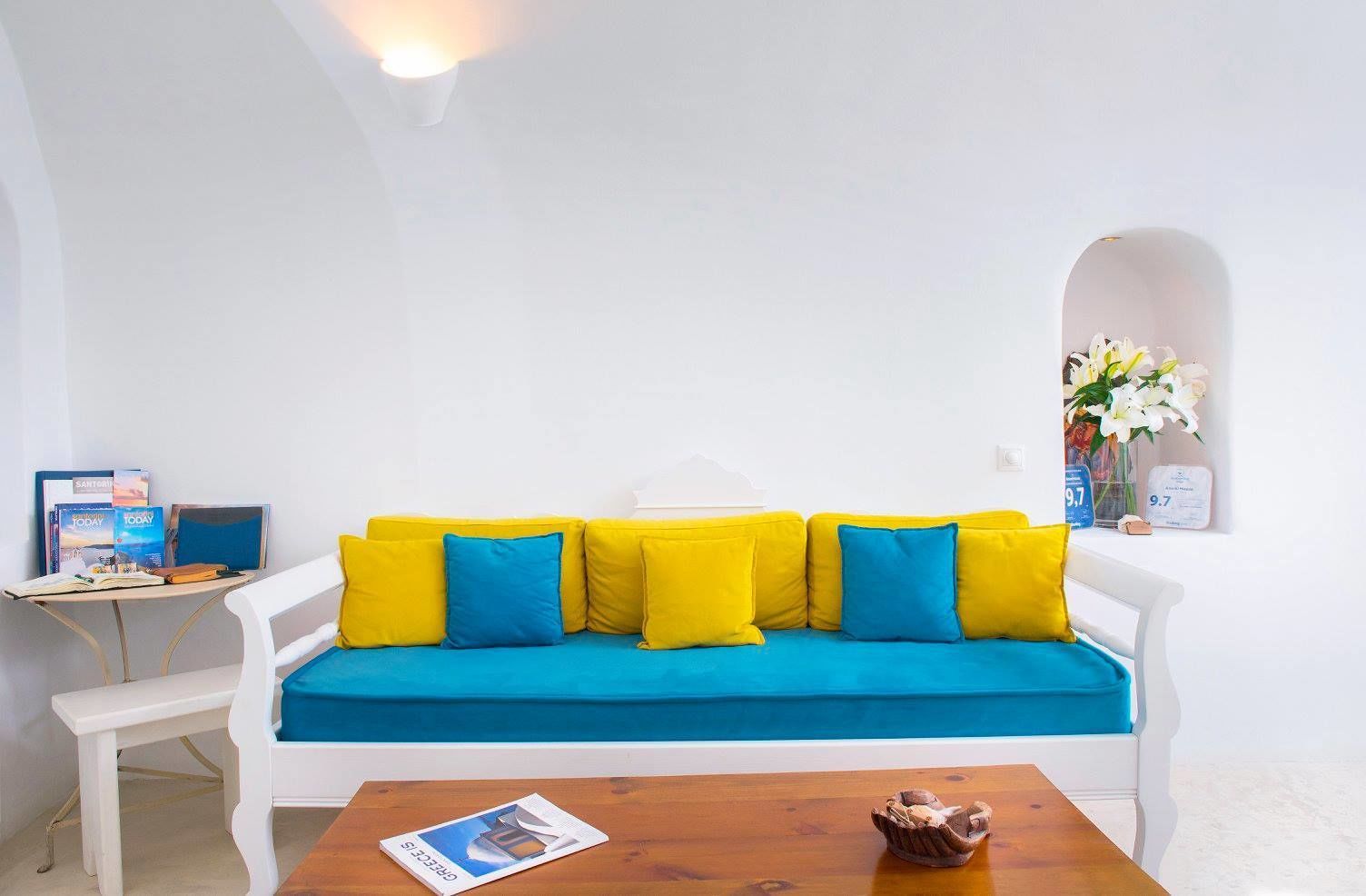 A white sofa with blue and yellow cushions, a wooden table with a magazine and a decorative item, and a small desk with books and a lamp.