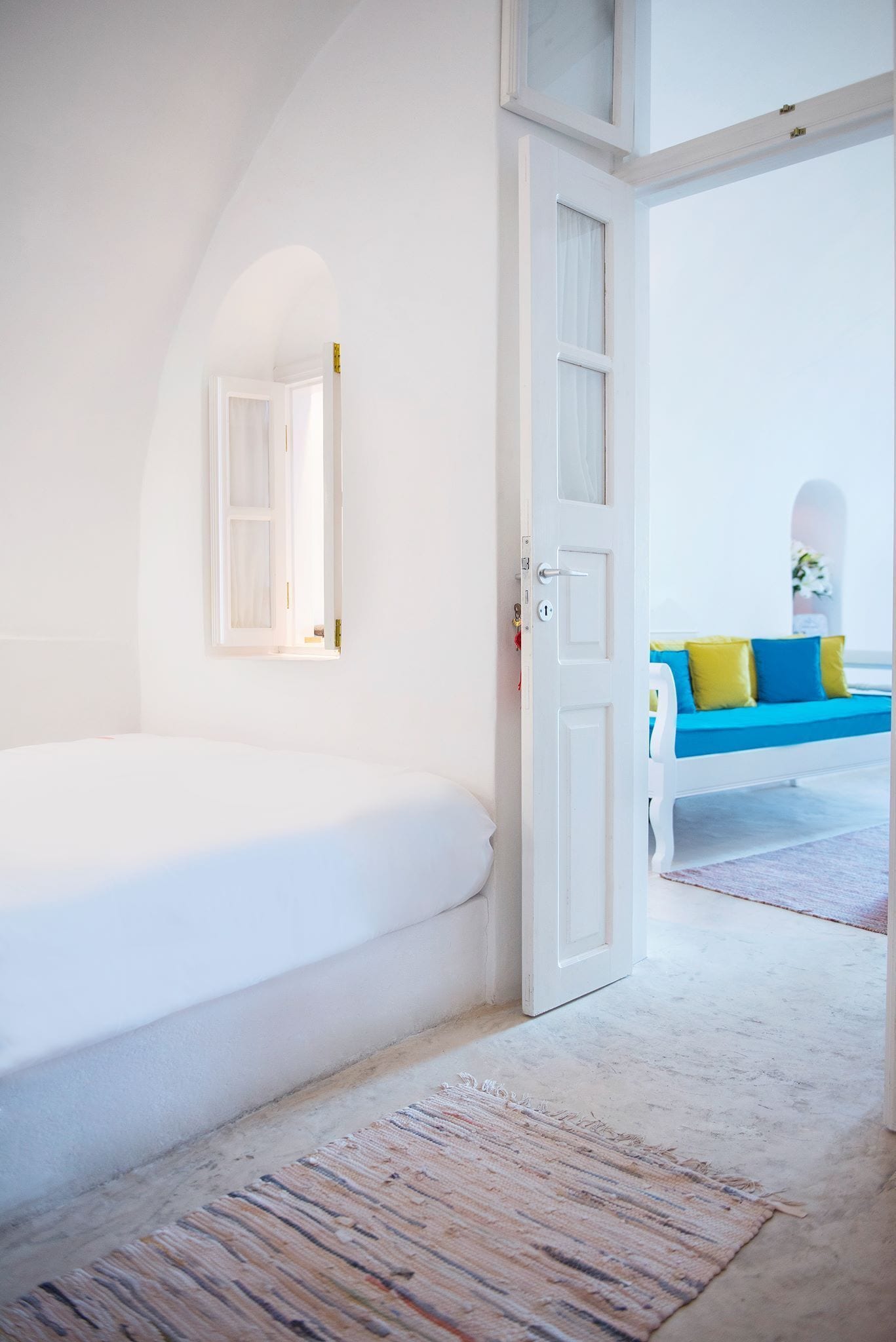 A bedroom with a white bed, a door leading to another room, and a view of a blue sofa with yellow pillows.