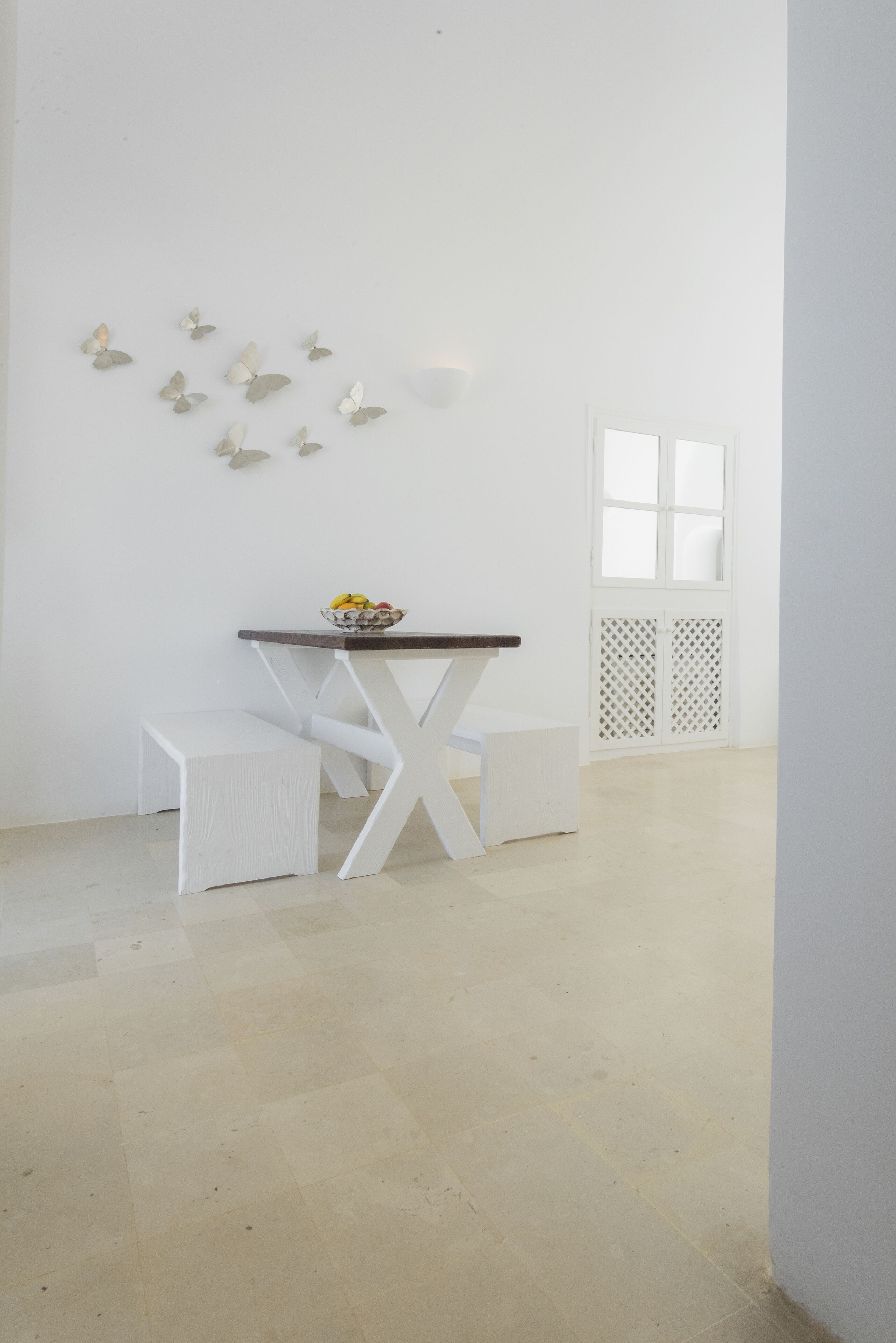 Un comedor minimalista con una mesa y banco blancos, un cuenco de fruta y decoración de mariposas en la pared.