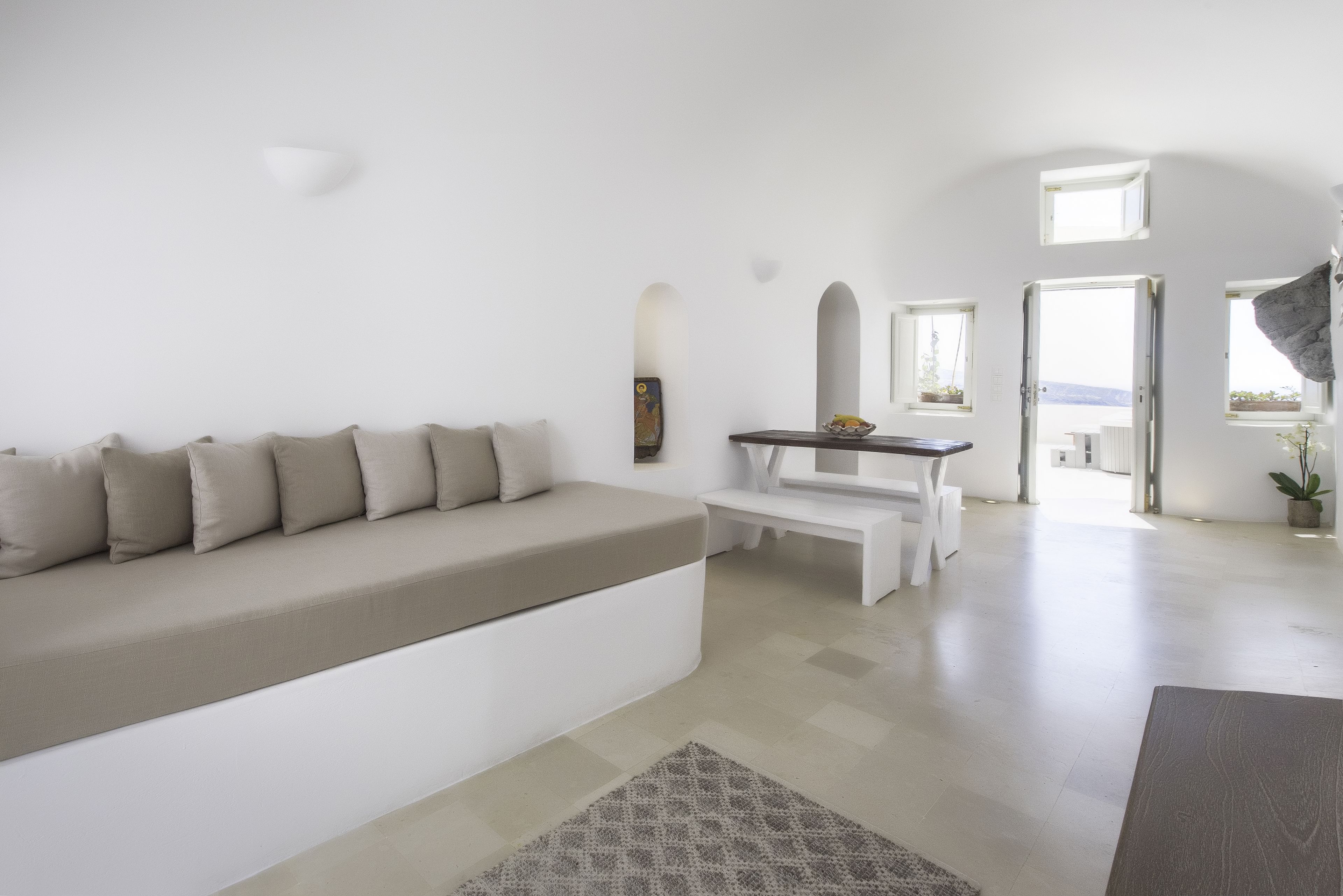 A modern living room with a sofa, a small table, and a bench. There are multiple windows and a view of the outdoors.