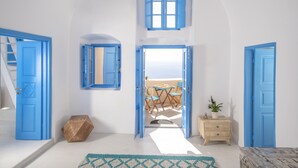 A bright room with blue doors, a wooden chest, a small table with chairs, and a view of the sea through an open door. - Anemi House & Villas (Santorini)