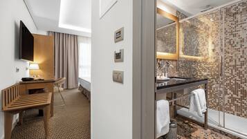 Habitació d'hotel amb escriptori de fusta, televisió de pantalla plana, bany modern amb dutxa de vidre i una catifes amb dibuixos.