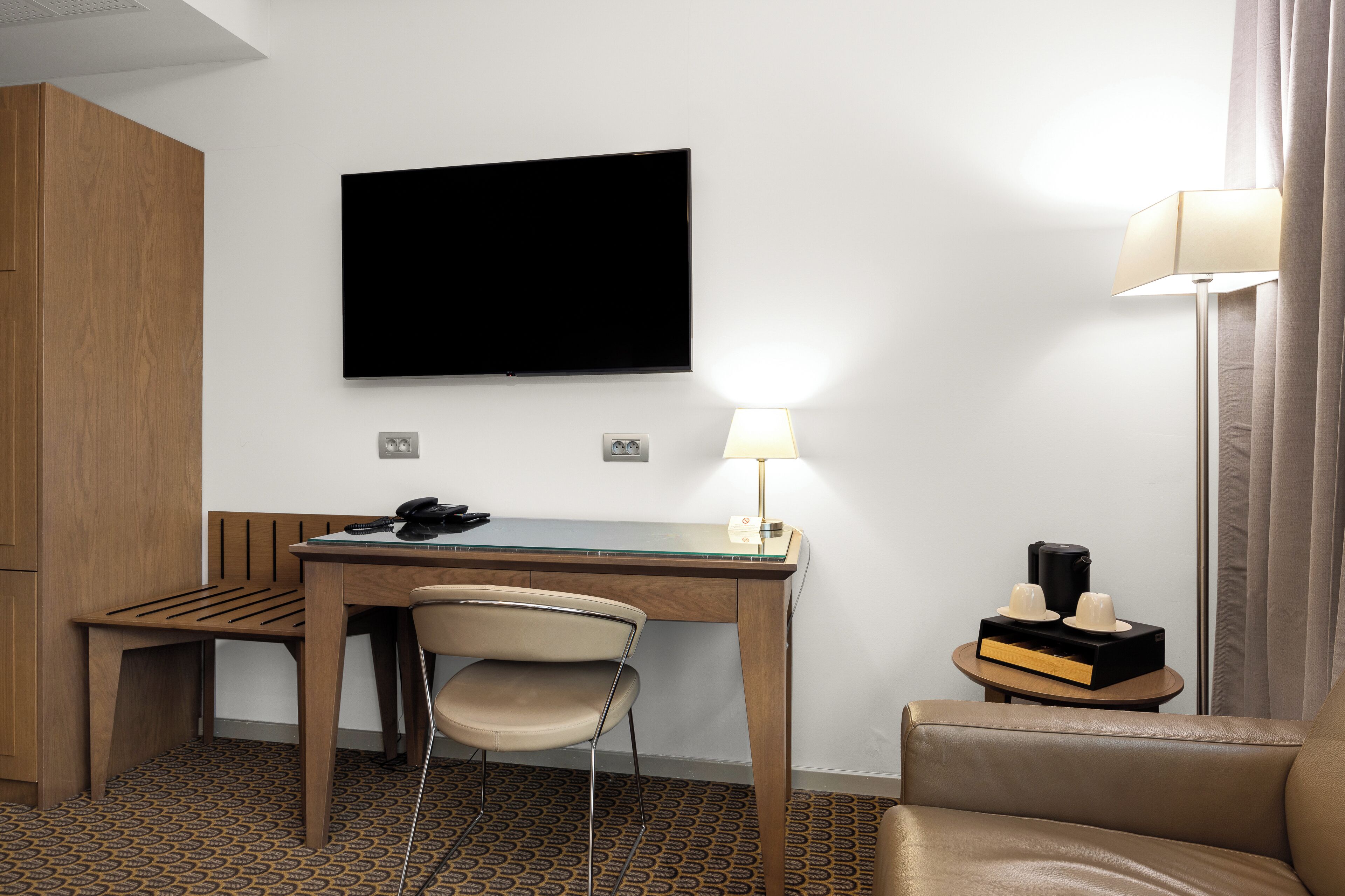 A hotel room with a wooden desk, a chair, a television mounted on the wall, a lamp, and a small table with a coffee maker and cups.