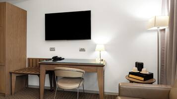 A hotel room with a wooden desk, a chair, a television mounted on the wall, a lamp, and a small table with a coffee maker and cups.