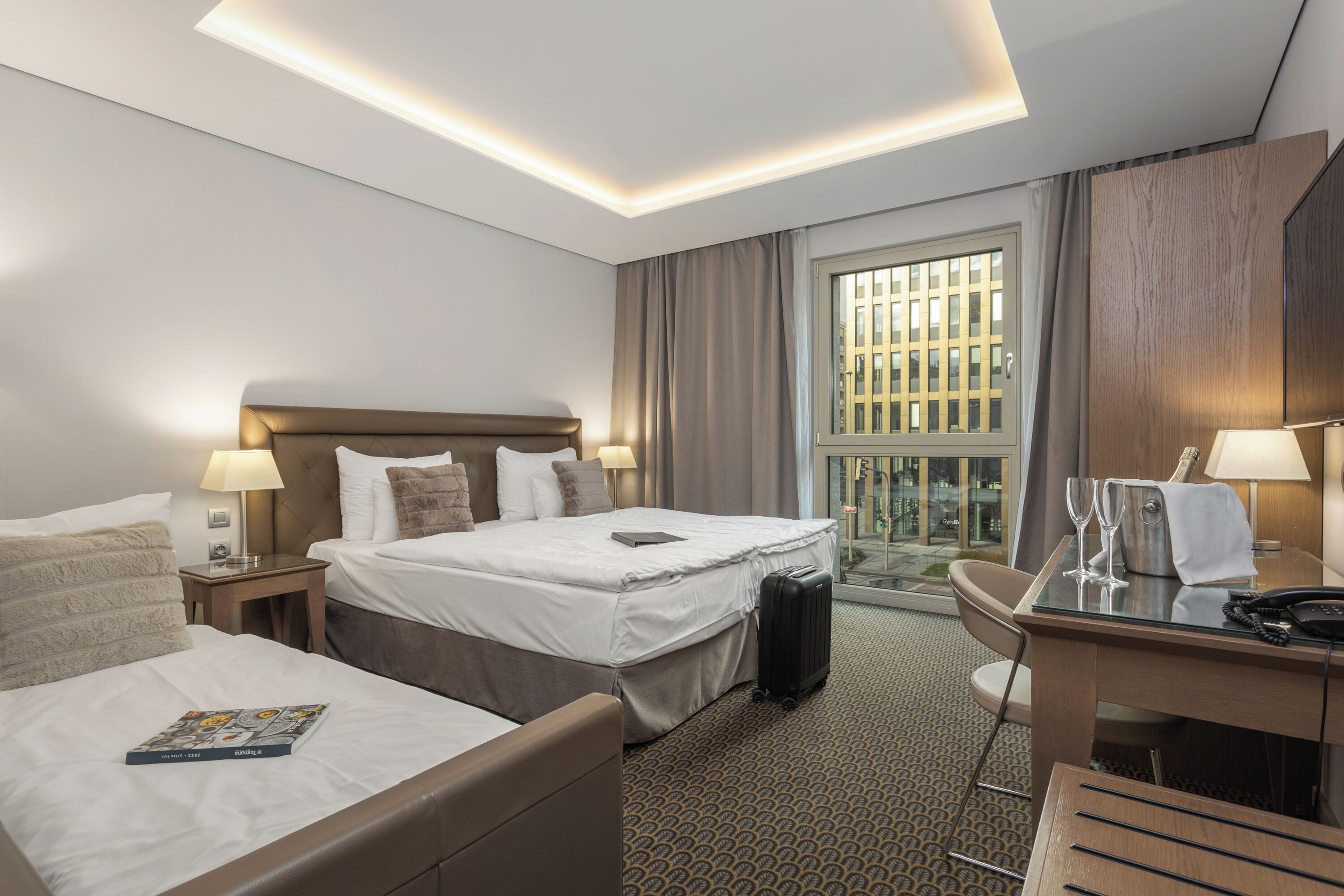 Habitación de hotel con dos camas, un escritorio, una silla, una maleta y ventana con vistas a edificios.