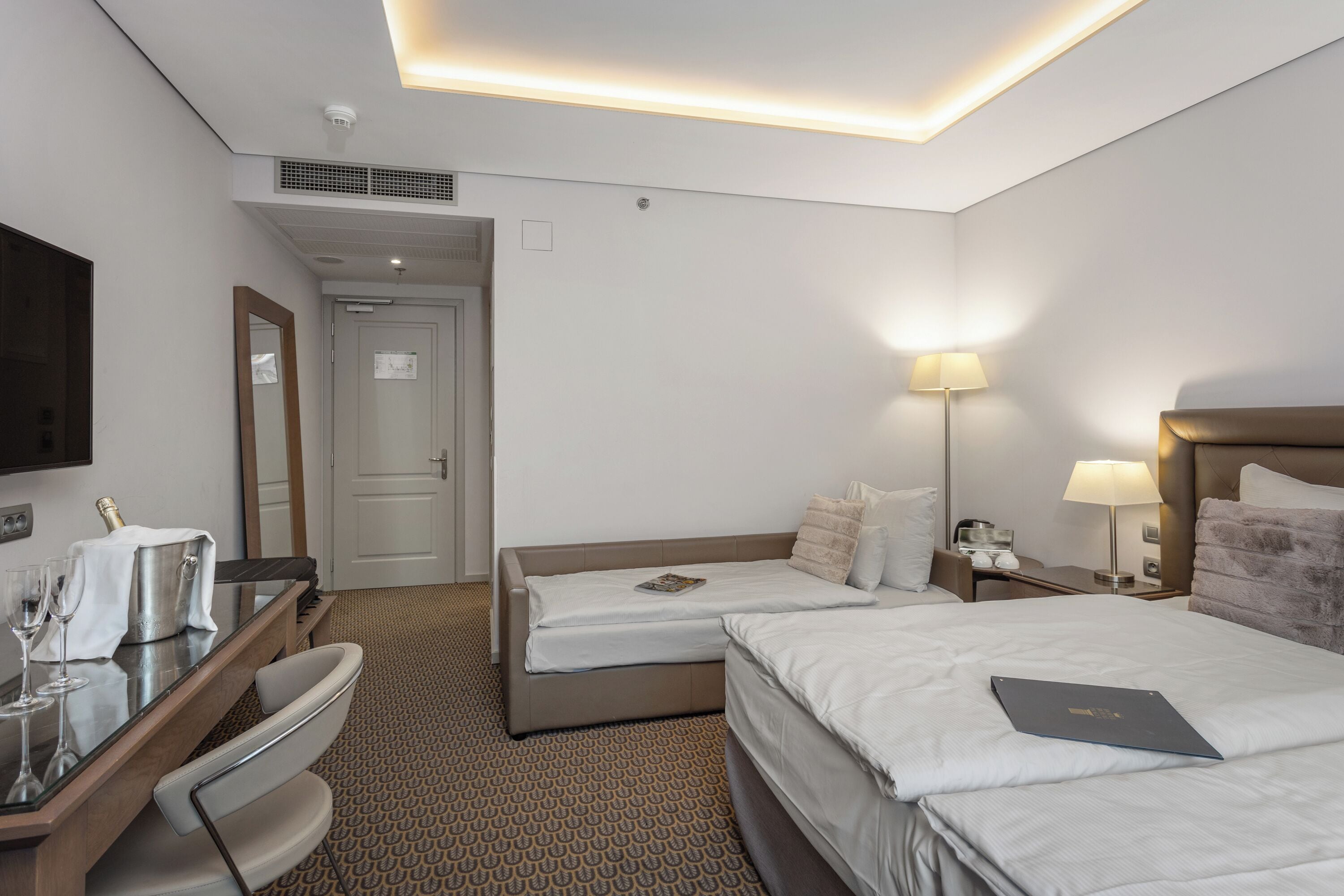 A hotel room with a bed, a desk with a chair, a lamp, and a minibar.