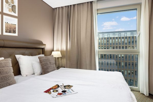 Una habitación de hotel con una cama grande, cabecero, ventana con vista a un edificio y una revista sobre la cama.