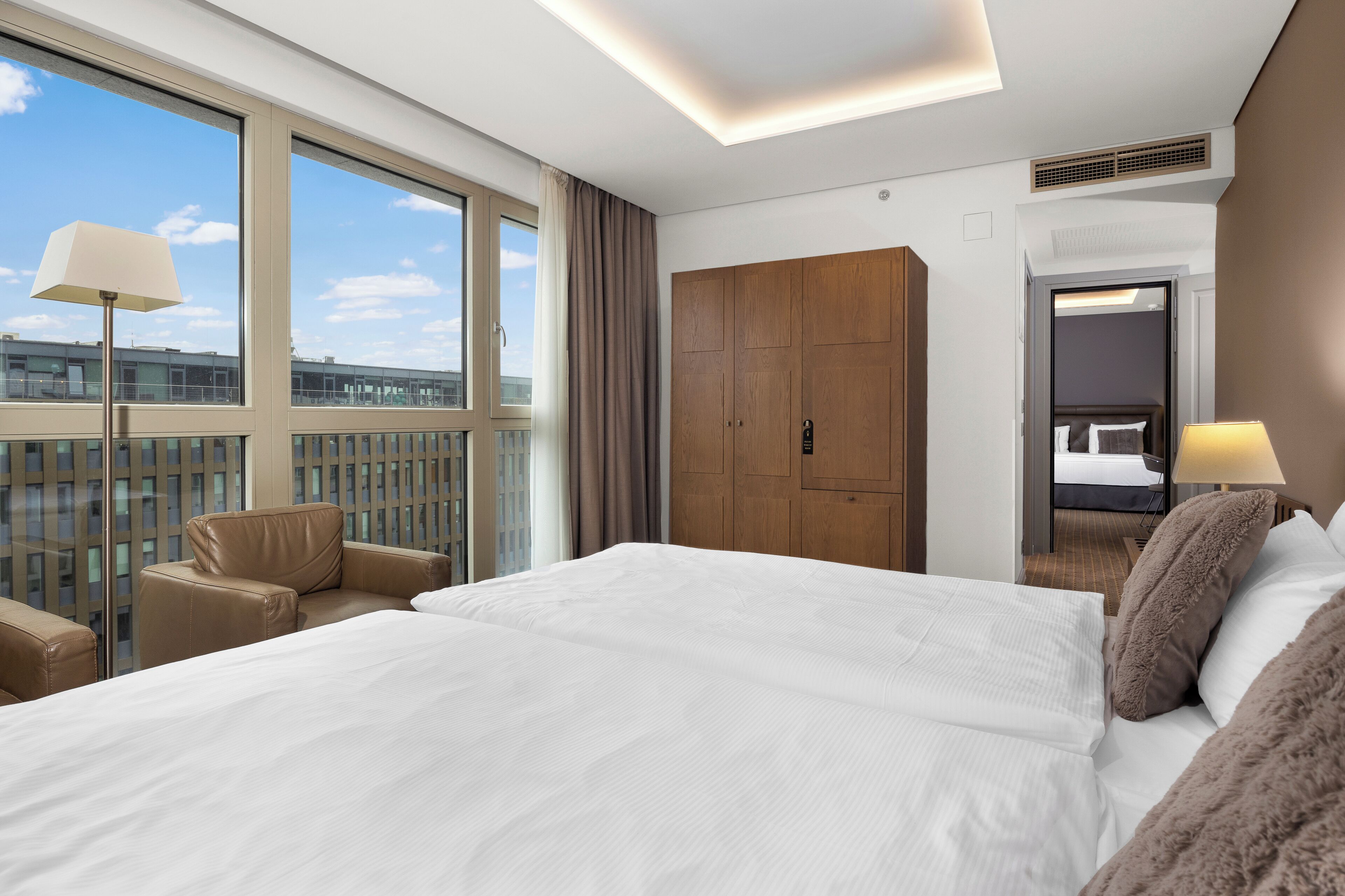 A hotel room with a large bed, a wooden wardrobe, a seating area with a chair, and a view of the city through large windows.