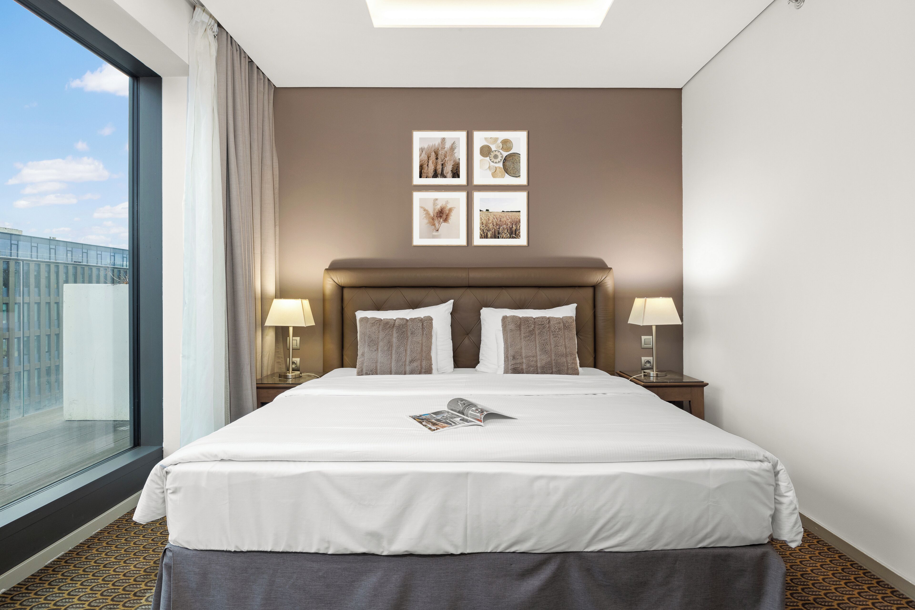 A hotel room with a large bed, two bedside lamps, a view of the city, and four framed pictures on the wall.