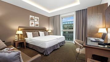 A hotel room with a large bed, a desk with a laptop, a chair, and a view of the city through the window.