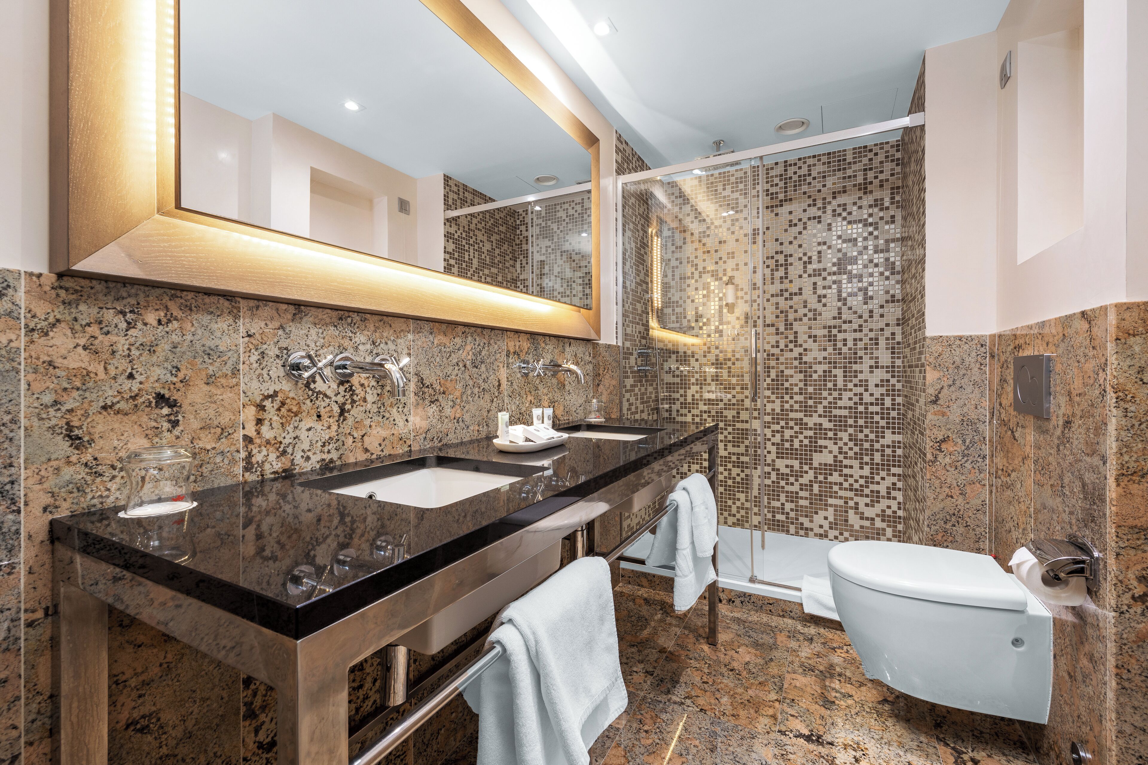 Un baño moderno con encimeras de granito, lavabo doble, cabina de ducha de cristal y un inodoro.