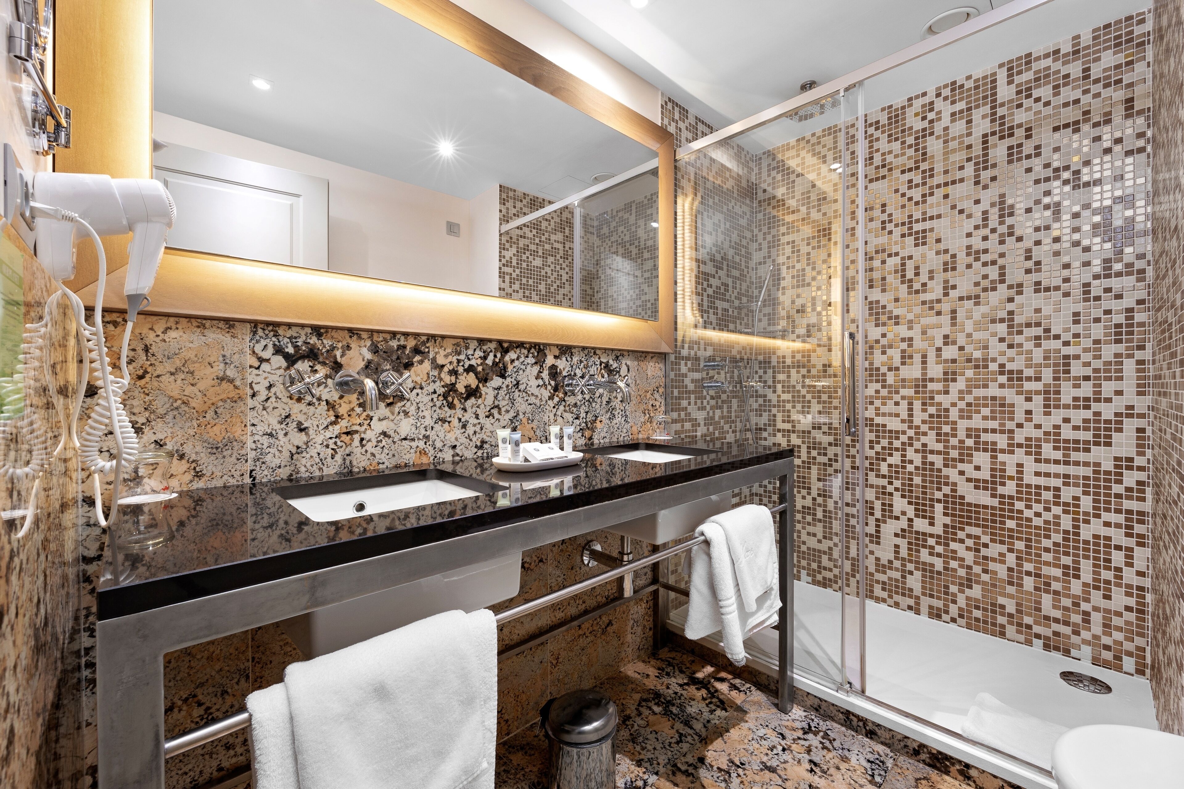 A modern bathroom with a granite countertop, two sinks, a large mirror, and a shower area with mosaic tiles.