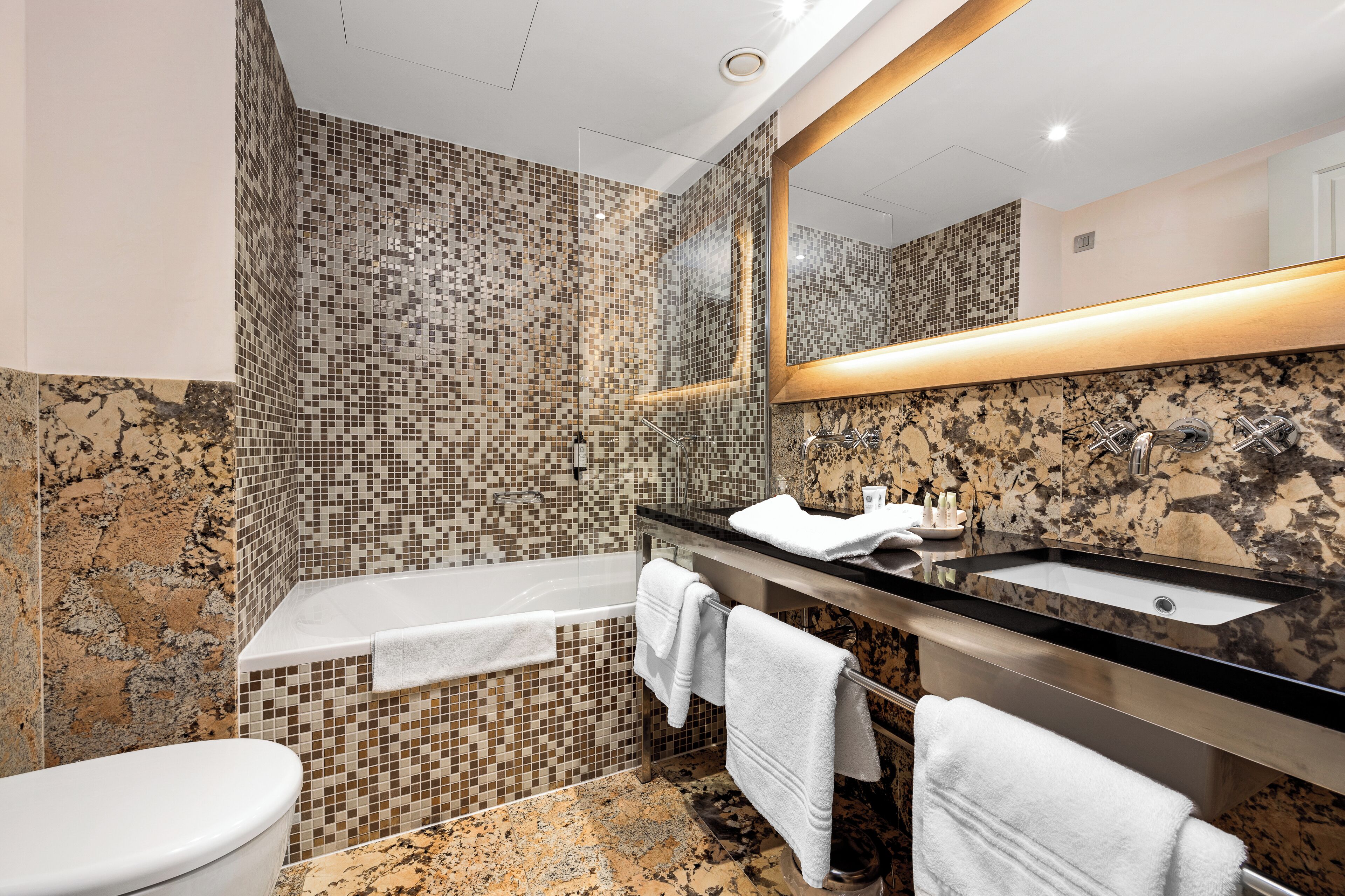 Une salle de bain moderne avec un mur recouvert de mosaïques, une baignoire, un lavabo doté d’un plan de travail noir et un grand miroir.