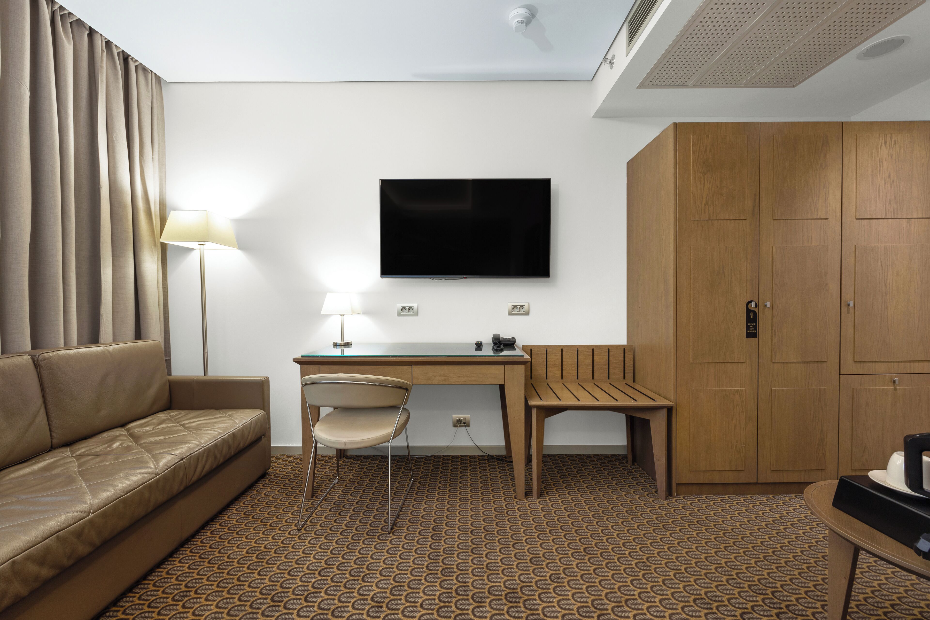Habitación de hotel con sofá, escritorio, silla, televisor, armario y alfombra estampada.