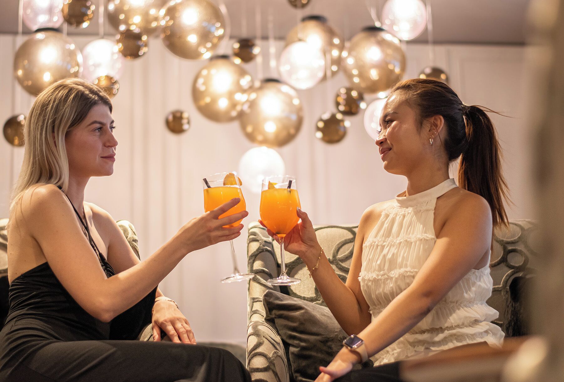 Dos personas sentadas en un sofá, sosteniendo bebidas, en una habitación con lámparas esféricas colgantes.