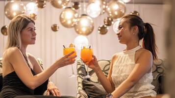 Dos personas sentadas en un sofá, sosteniendo bebidas, en una habitación con lámparas esféricas colgantes.