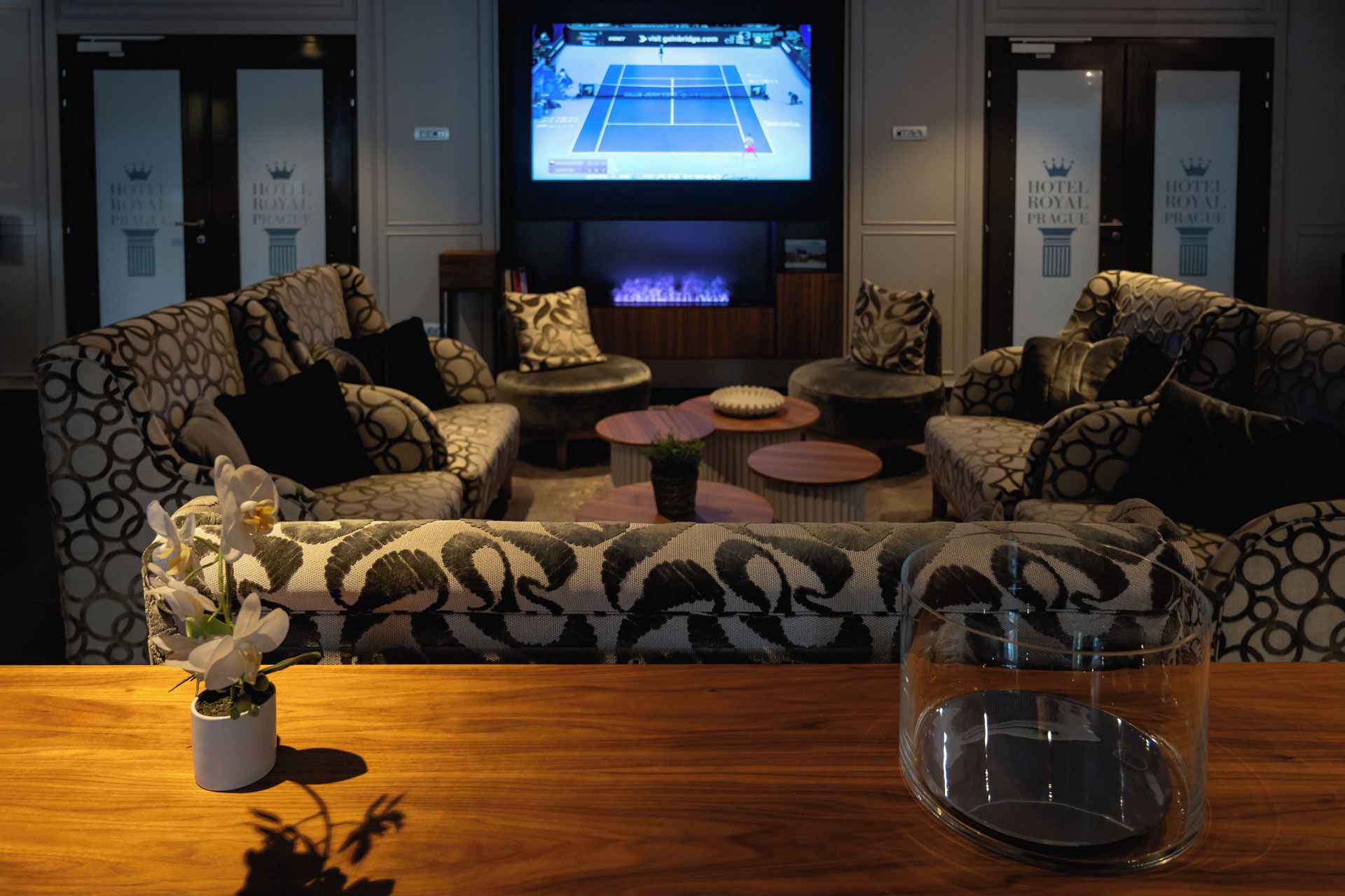 Un espace détente avec des canapés à motifs, un téléviseur diffusant un match de tennis et une table agrémentée d’un vase de fleurs.