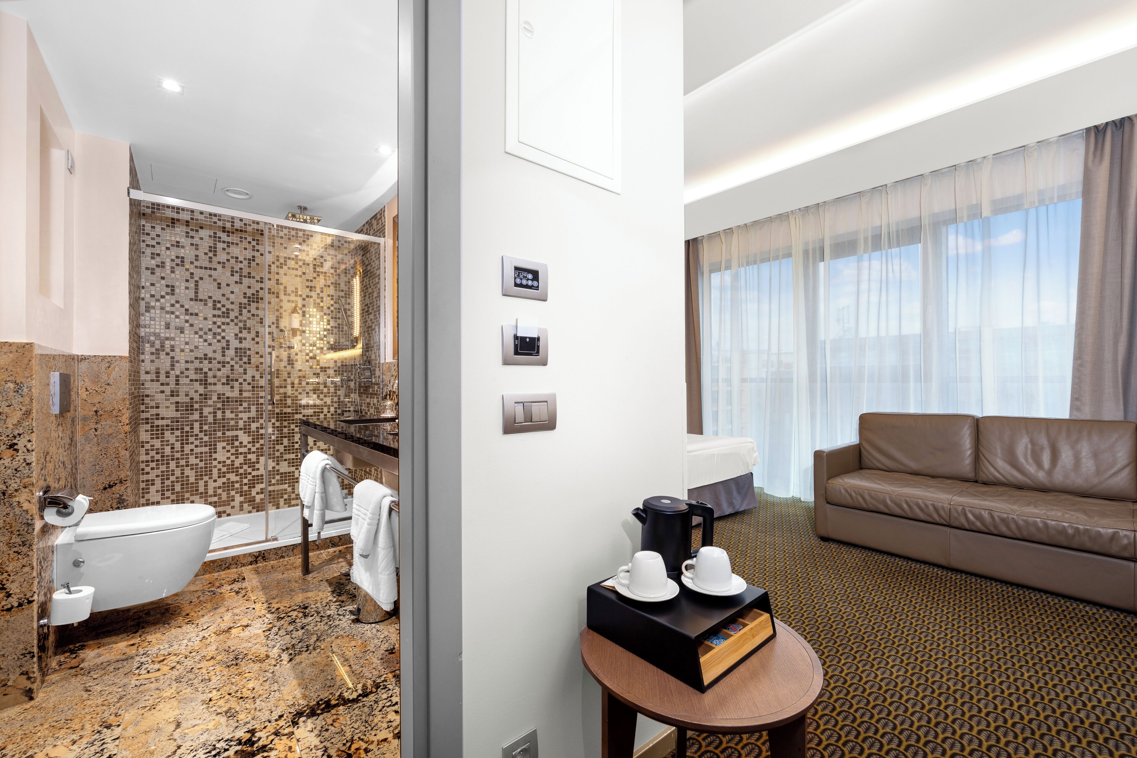 A modern hotel room with a bathroom featuring a mosaic tile wall, a white toilet, and a shower area. There is a brown leather sofa and a small table with a tray holding cups and a book.