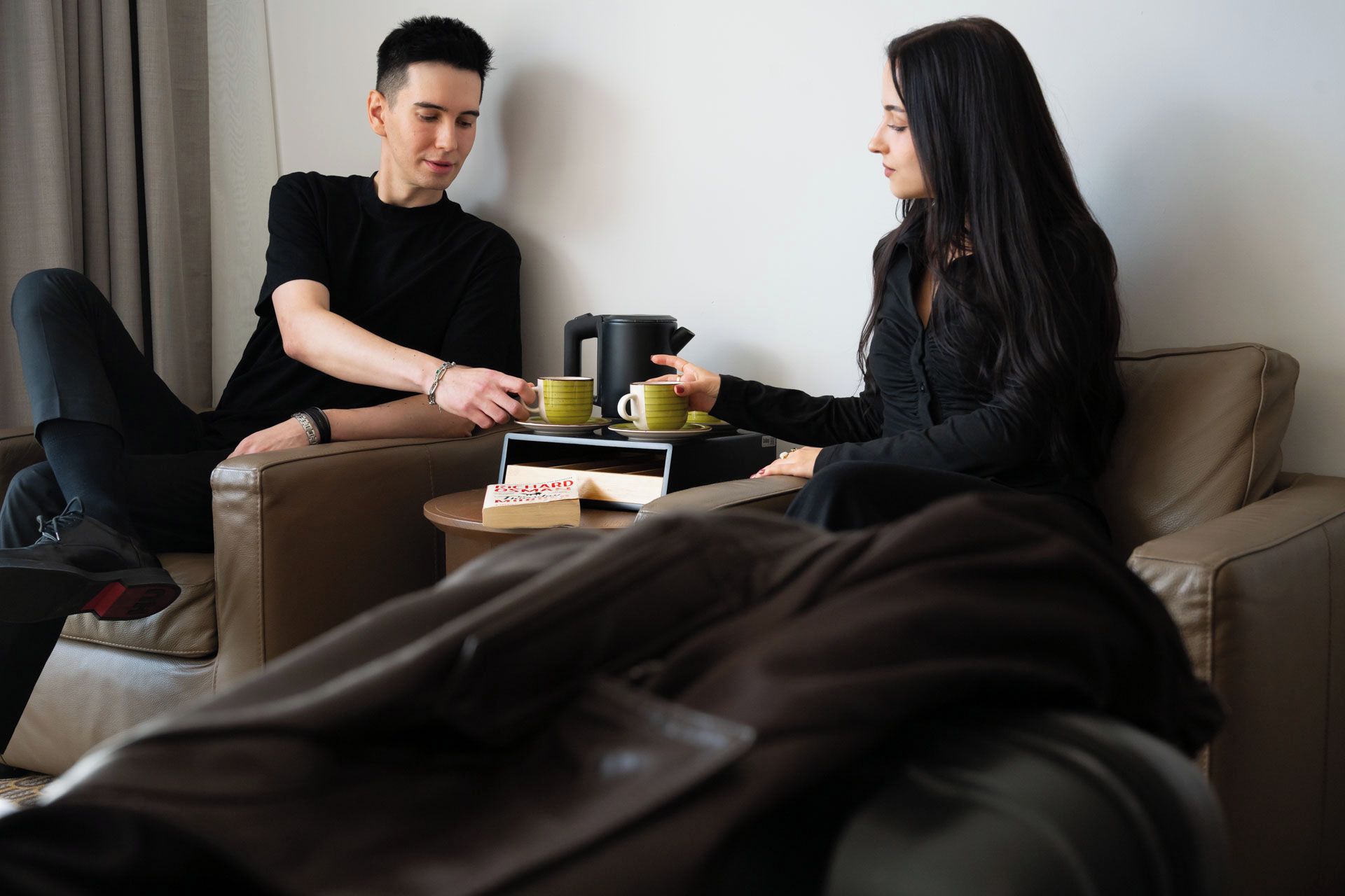Duas pessoas sentadas em um sofá, com uma cafeteira e um livro sobre uma mesinha.