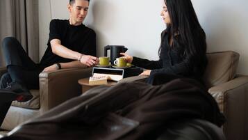 Duas pessoas sentadas em um sofá, com uma cafeteira e um livro sobre uma mesinha.
