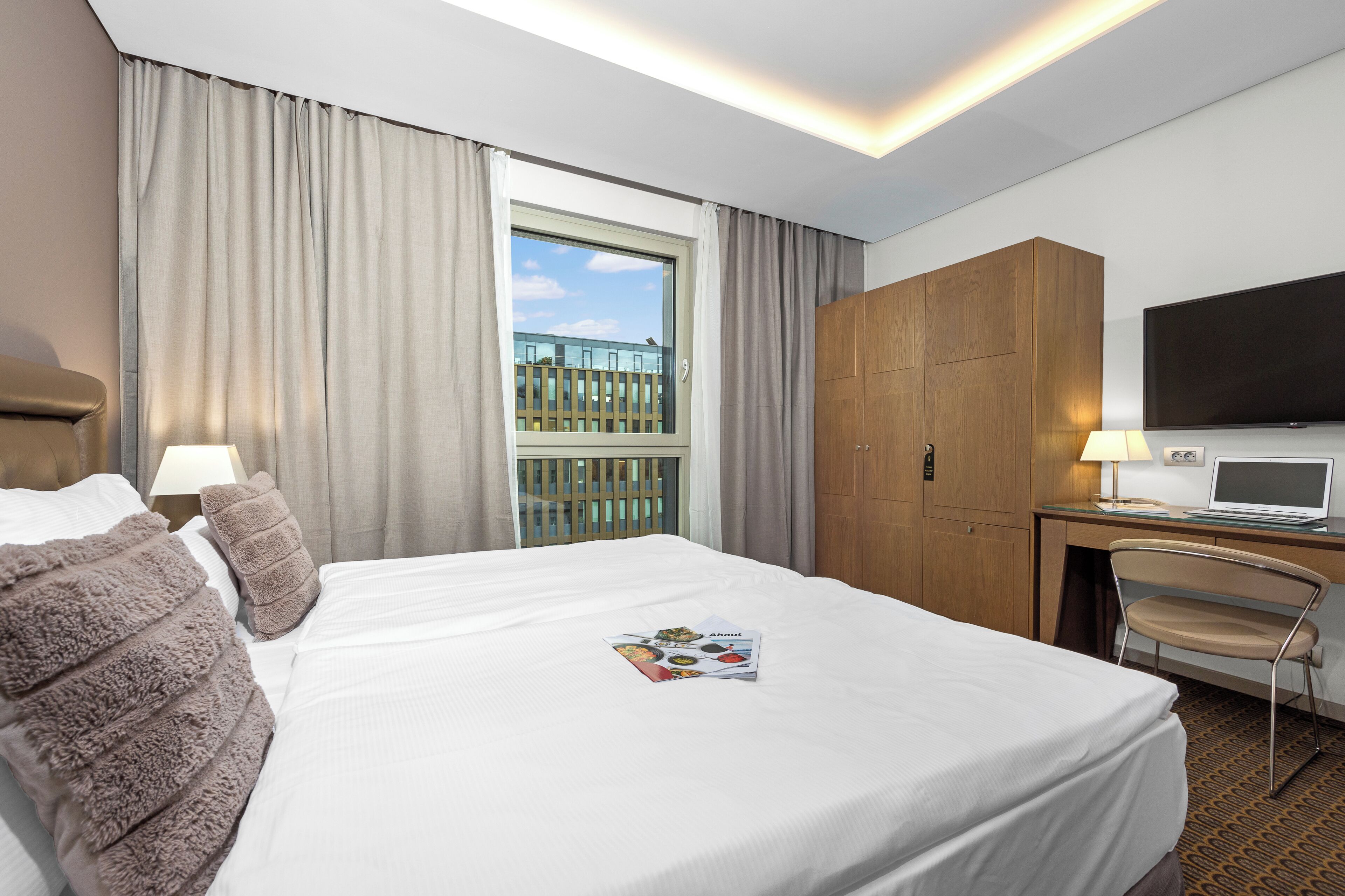 Et hotelværelse med en stor seng, et skrivebord med en bærbar computer, et fjernsyn og et vindue med gardiner.