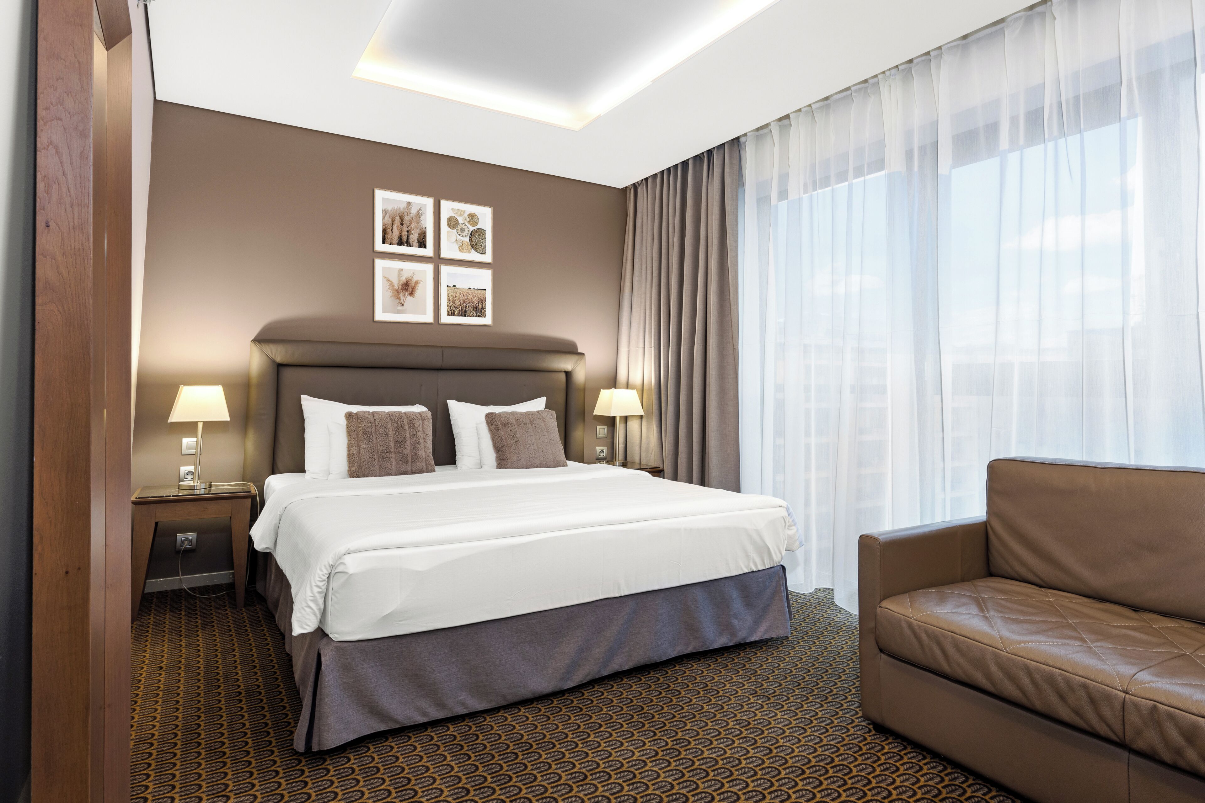 Una habitación de hotel con una cama grande, dos mesitas de noche con lámparas, un sofá marrón y una alfombra estampada.