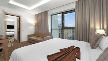 A modern hotel room with a large bed, a desk, a chair, and a view of the city through a large window.