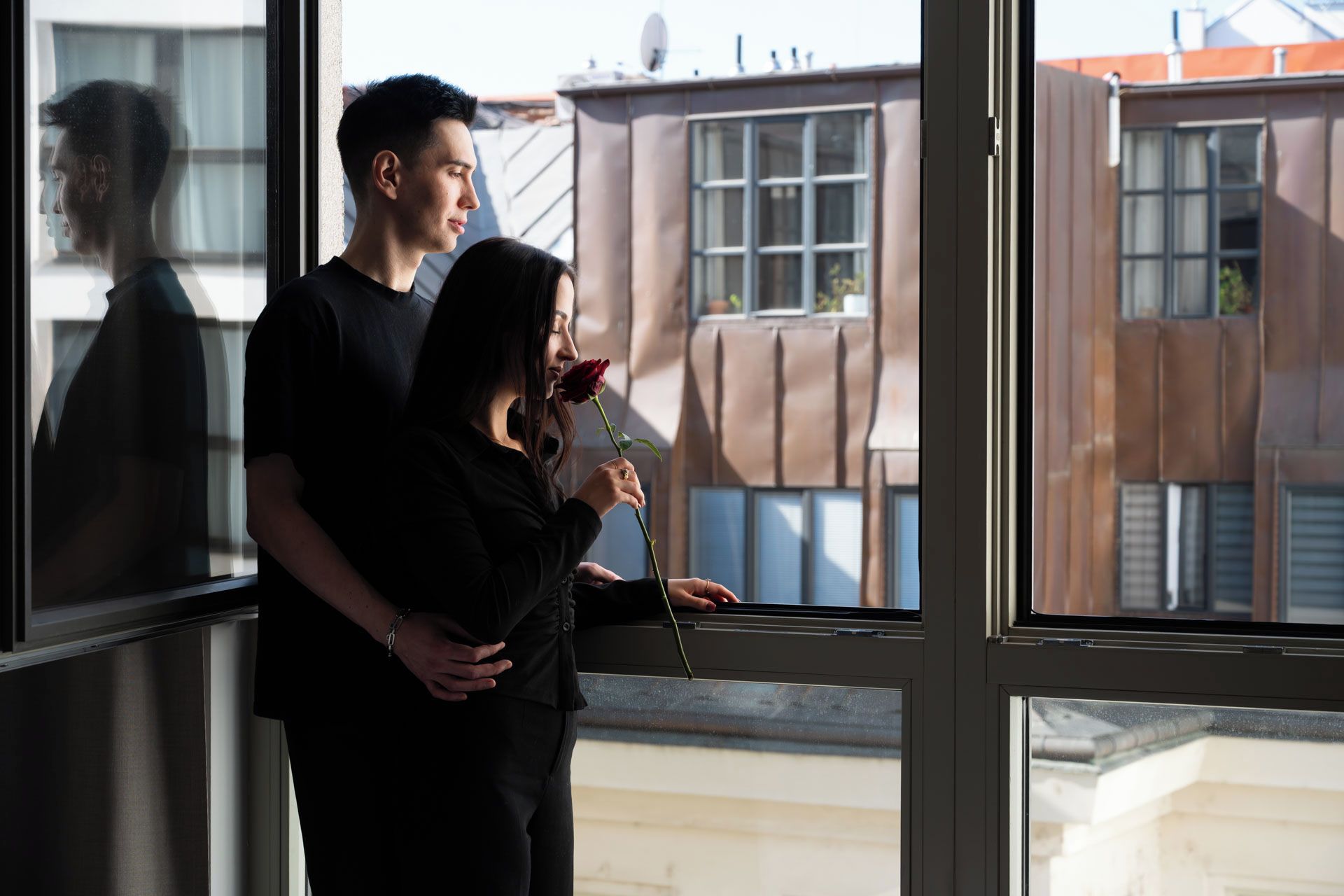 A couple standing by a window with a view of buildings outside.