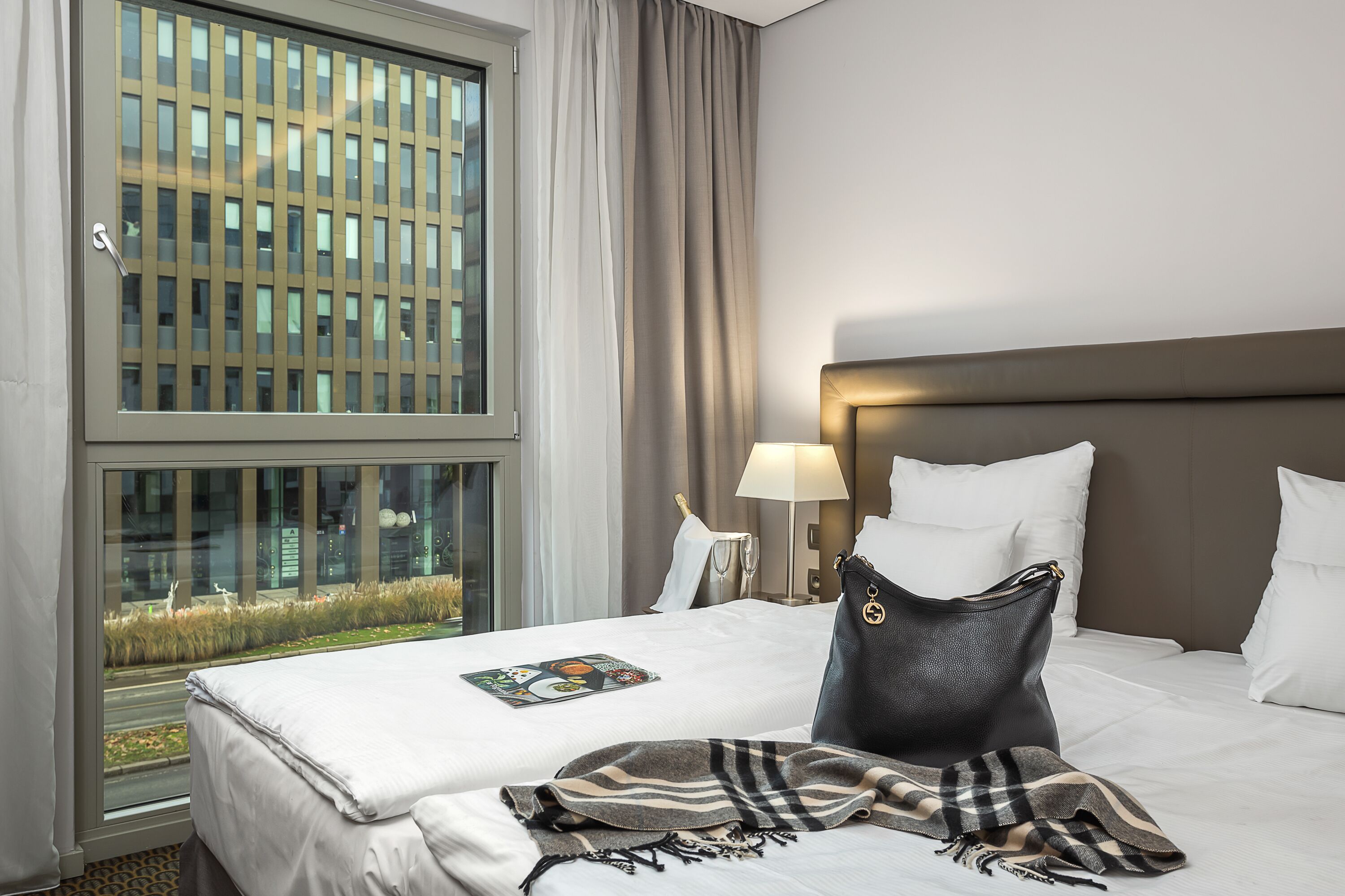 A hotel room with a large window, a bed with white linens, a black handbag, and a lamp on the nightstand.