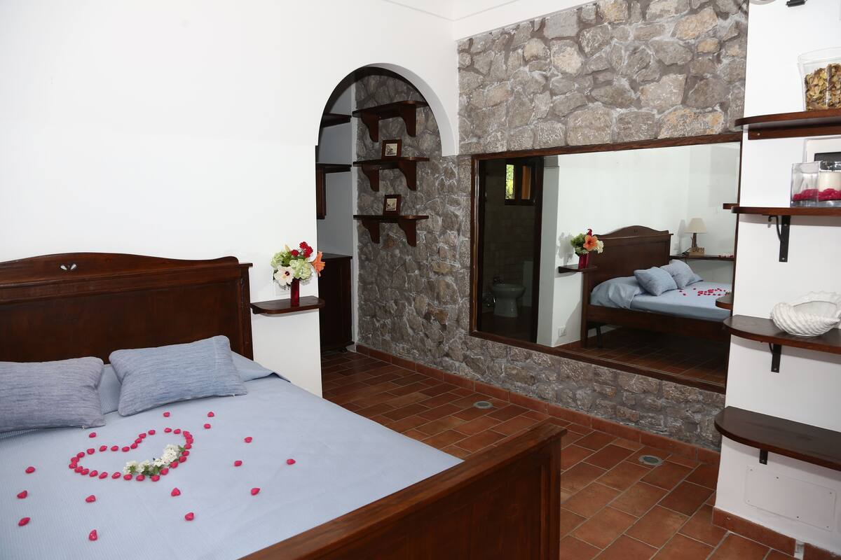 A bedroom with a stone wall, a wooden bed, a mirror, and shelves with decorative items.