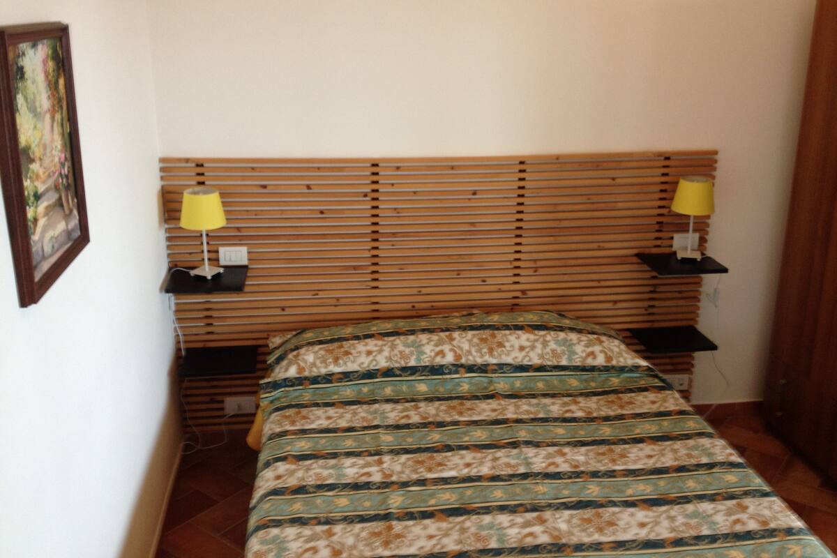 A small, neatly arranged bedroom with a wooden slatted headboard, two bedside tables with lamps, and a patterned bedspread.