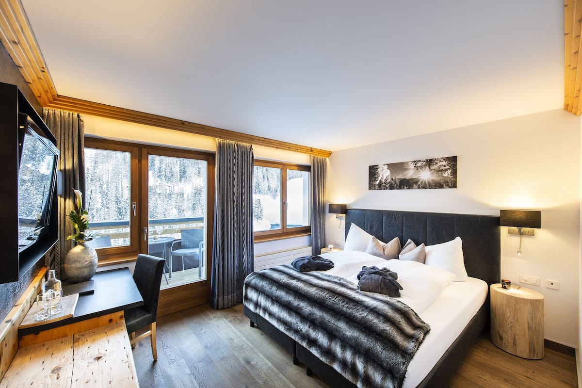 Habitación de hotel con cama, escritorio, silla, televisor, ventana con cortinas y luz instalada en la pared.