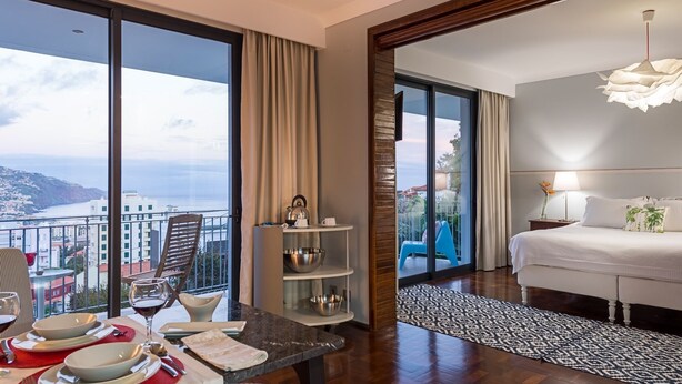 A hotel room with a balcony, a dining area with a table set for two, and a bed with a view of the city. - Belvedere Boutique - Adults Only (Funchal)